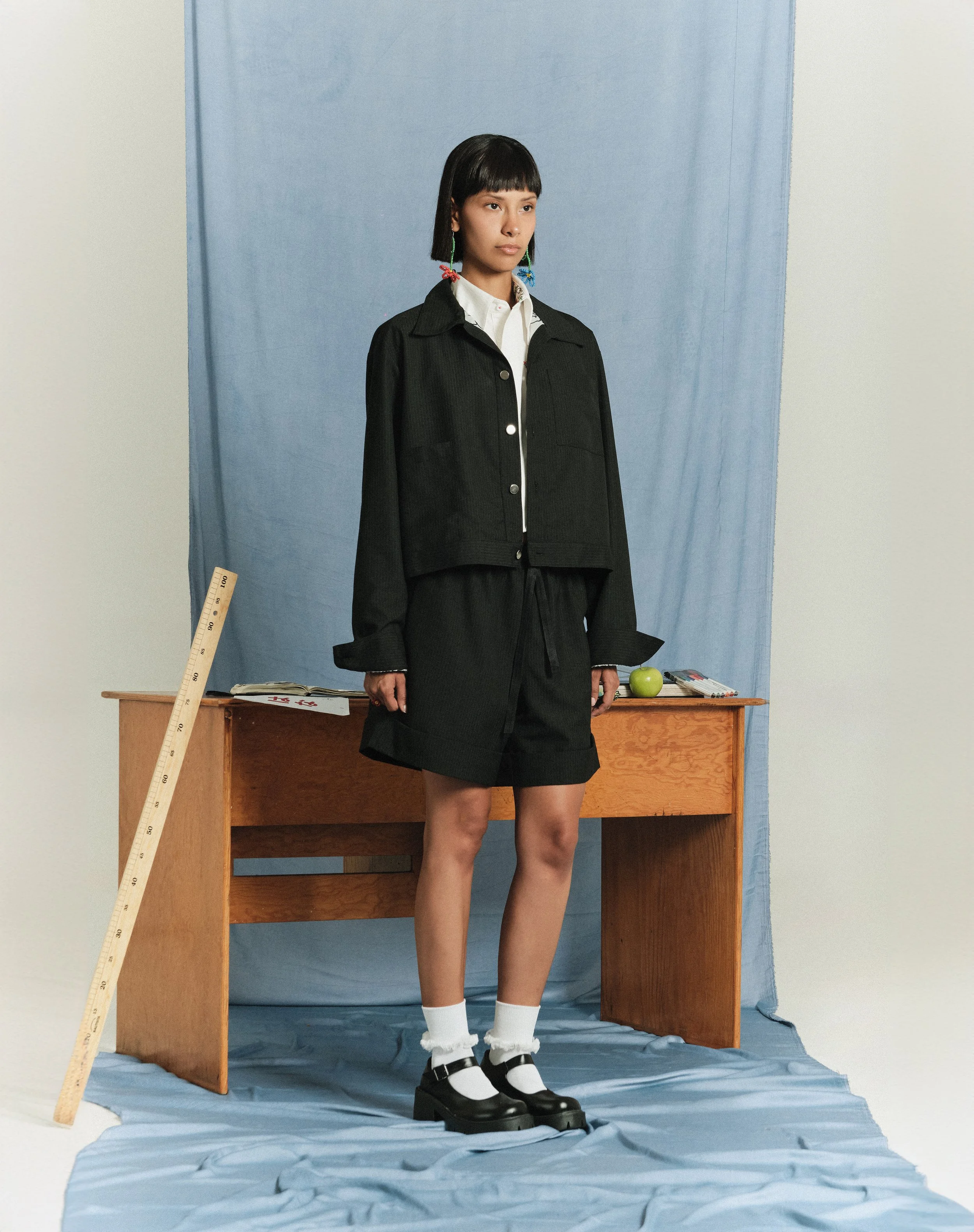 Joven mujer con corte de cabello recto y flequillo, vestida con chaqueta negra, camisa blanca, falda shorts negra, calcetas blancas y zapatos con hebillas negras, de pie frente a un escritorio de madera en un fondo de tela azul y pared blanca.