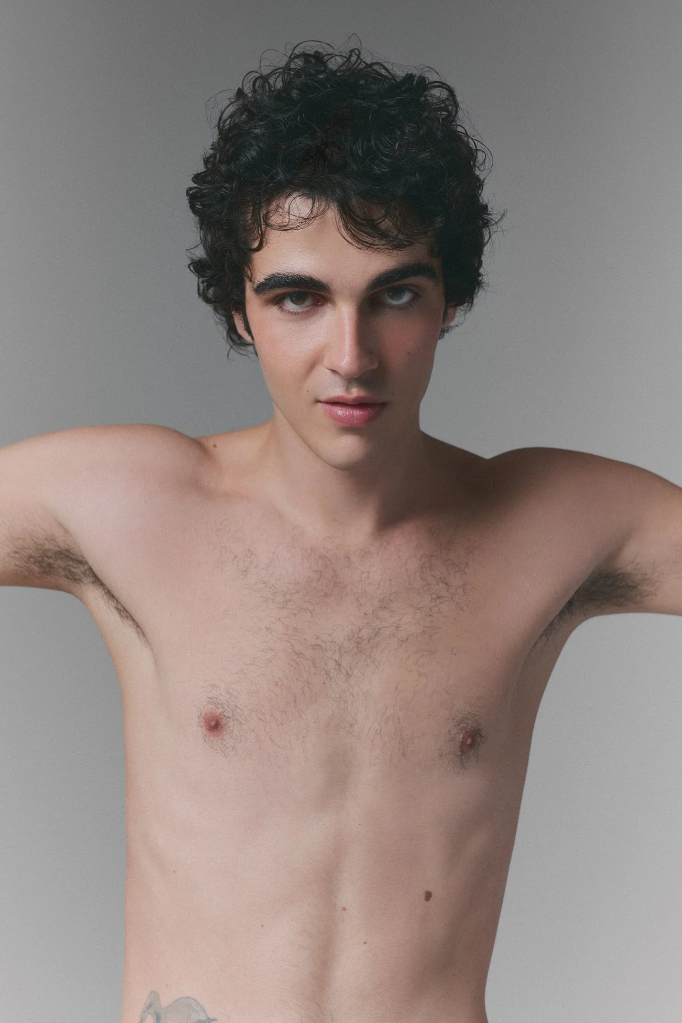 Hombre joven sin camisa, con cabello rizado oscuro, cejas gruesas, ojos azul claro, piel clara, en un fondo gris y con un tatuaje en el torso, mirando a la cámara.