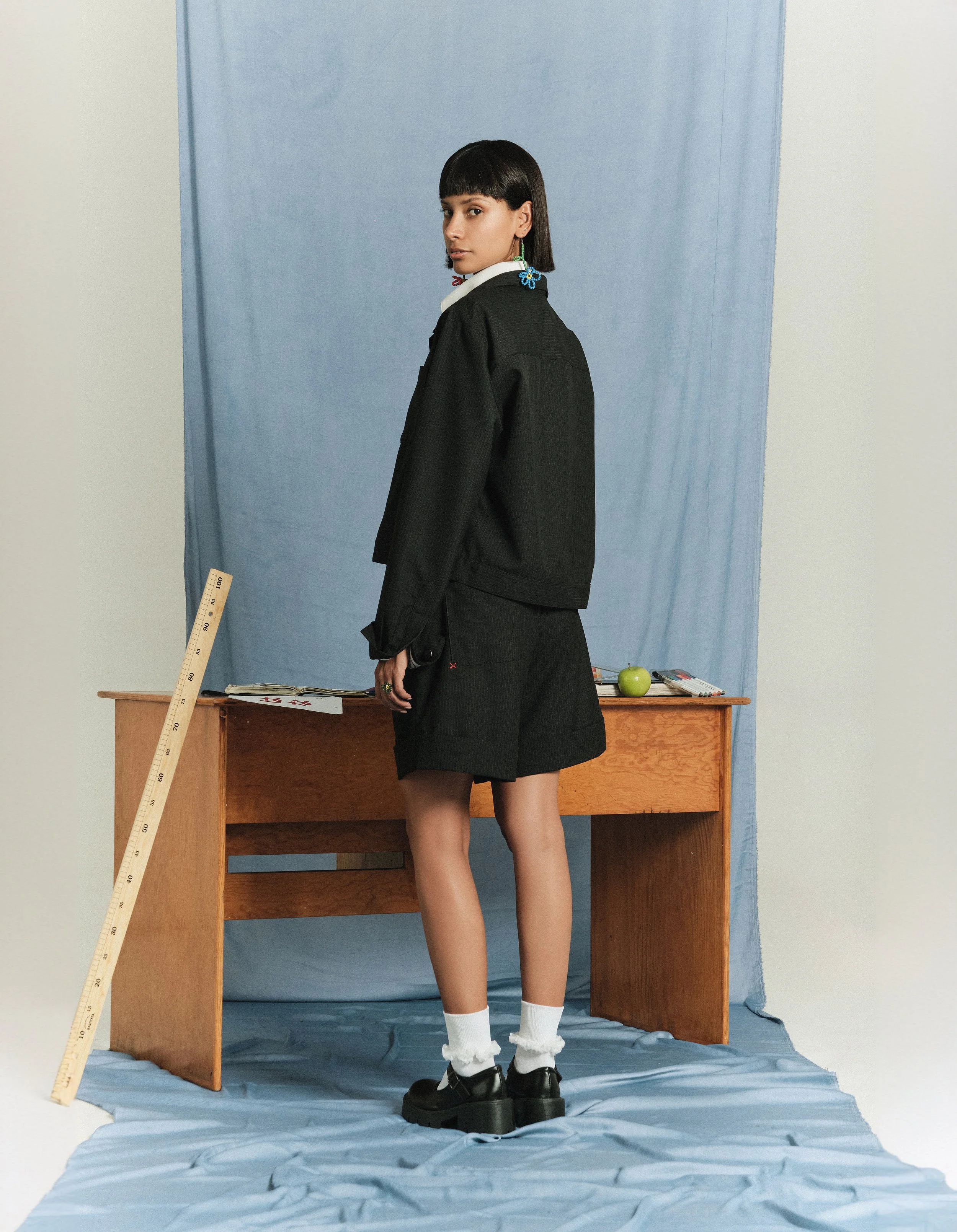 Juvenil mujer en ropa oscura de pie frente a un escritorio de madera con objetos y un balón en la mesa, en un fondo con tela azul y pared blanca.