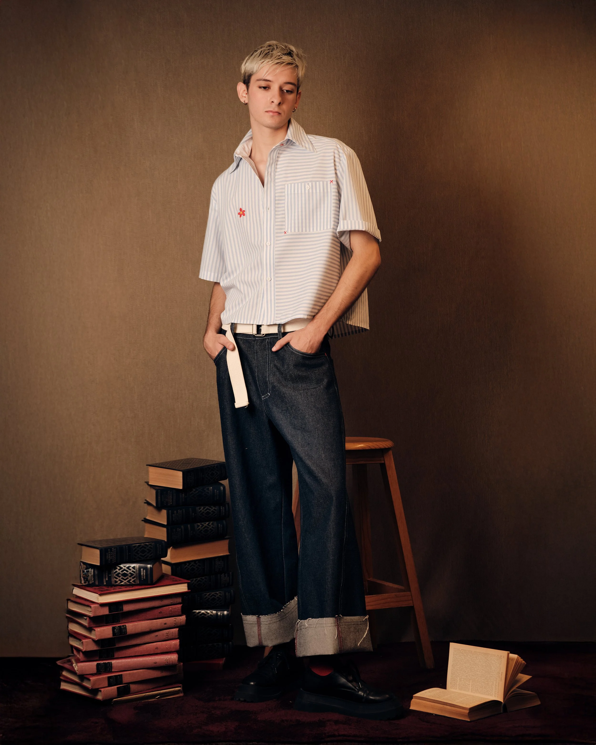 Una persona con cabello rubio corto y peinado hacia un lado, viste una camisa de rayas blancas y azules con bolsillos y detalles rojos, pantalones de mezclilla oscuros con los dobladillos doblados y zapatos negros, en un ambiente con fondo beige y va