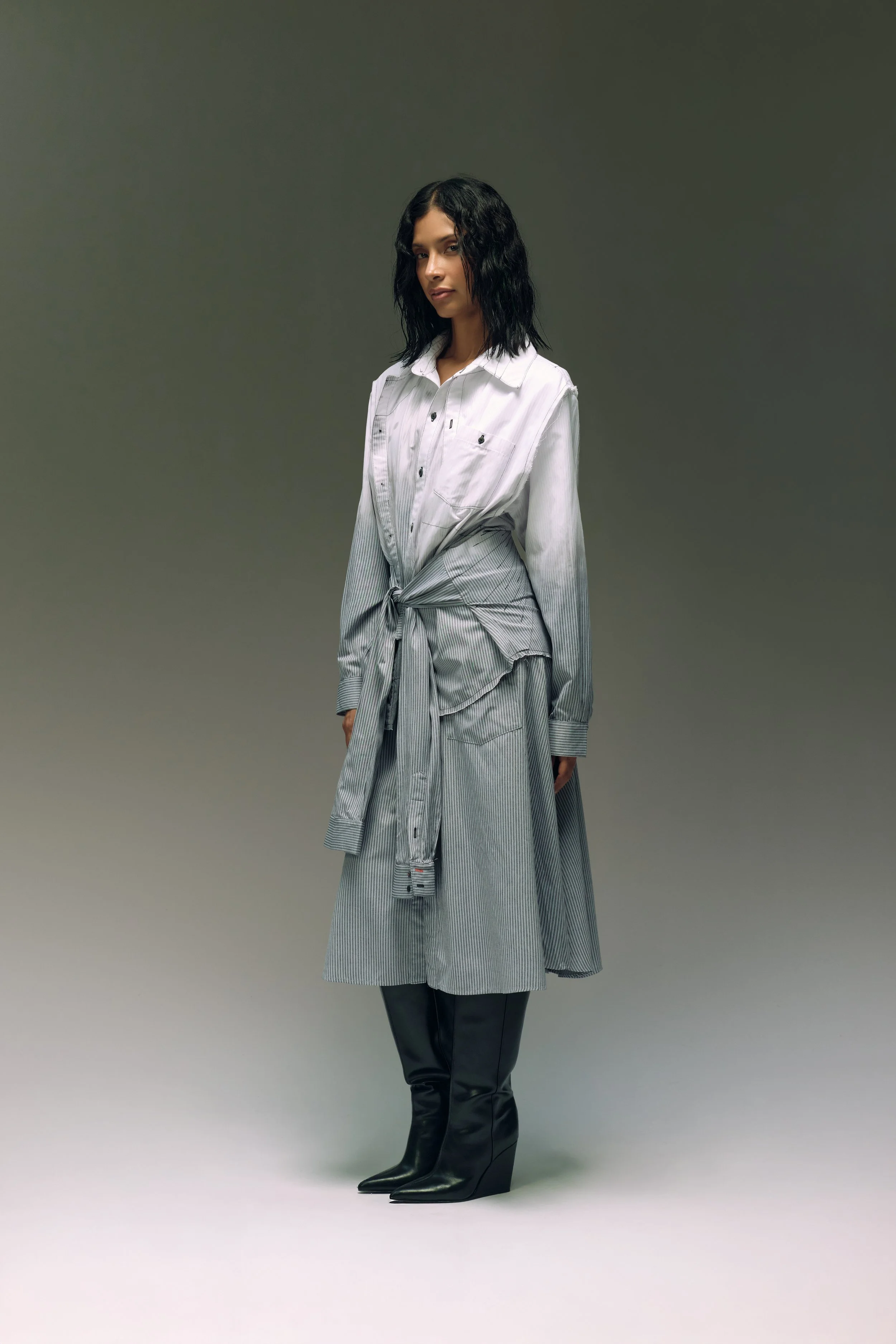 Mujer con blusa de mezclilla y falda larga a rayas, con botas negras de tacón alto en un fondo gris uniforme.