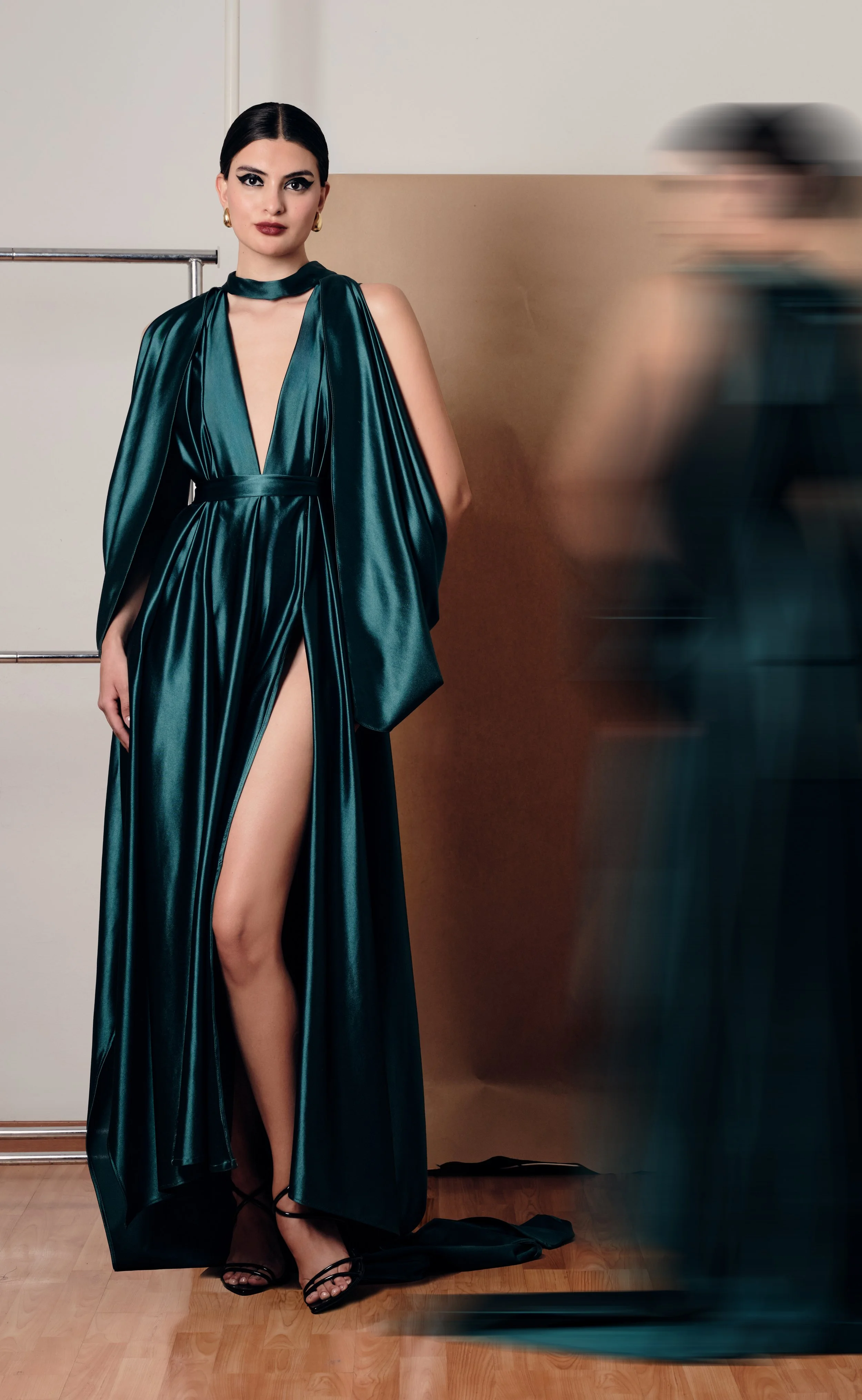 Mujer con vestido largo de satén azul oscuro y polleras abiertas, zapatos de tacón negro, peinado liso y peinada, accesorios dorados, en un ambiente sencillo, con una persona en movimiento borroso a la derecha.