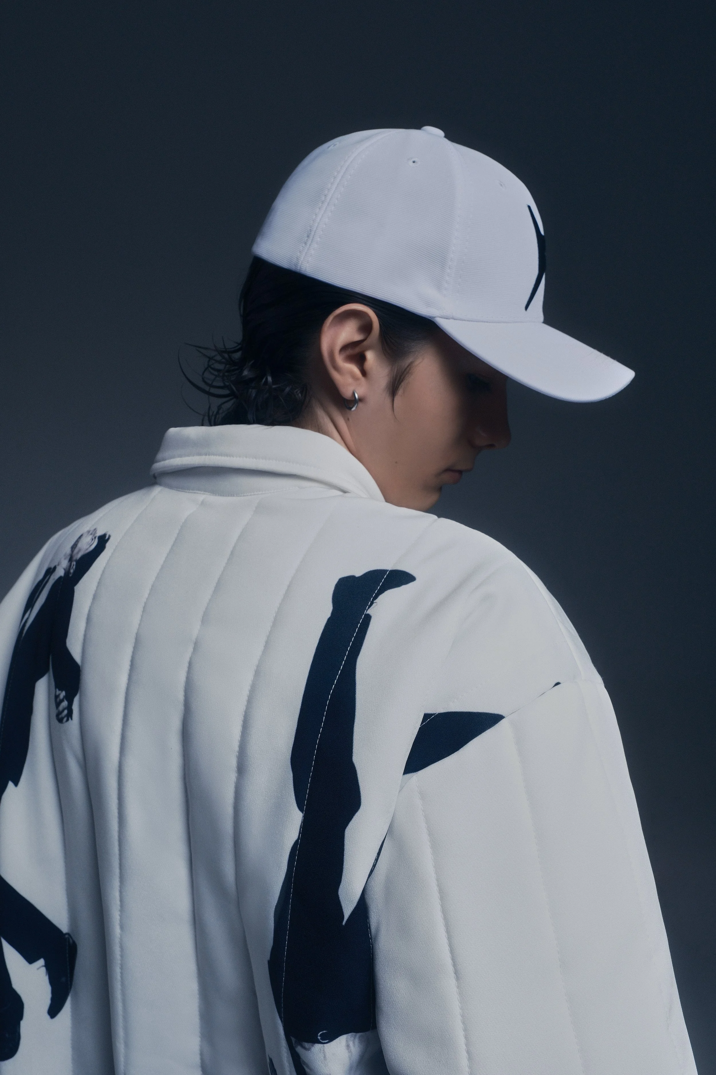 Persona con gorra blanca y chaqueta blanca con detalles en negro, mirando hacia abajo en un fondo oscuro.