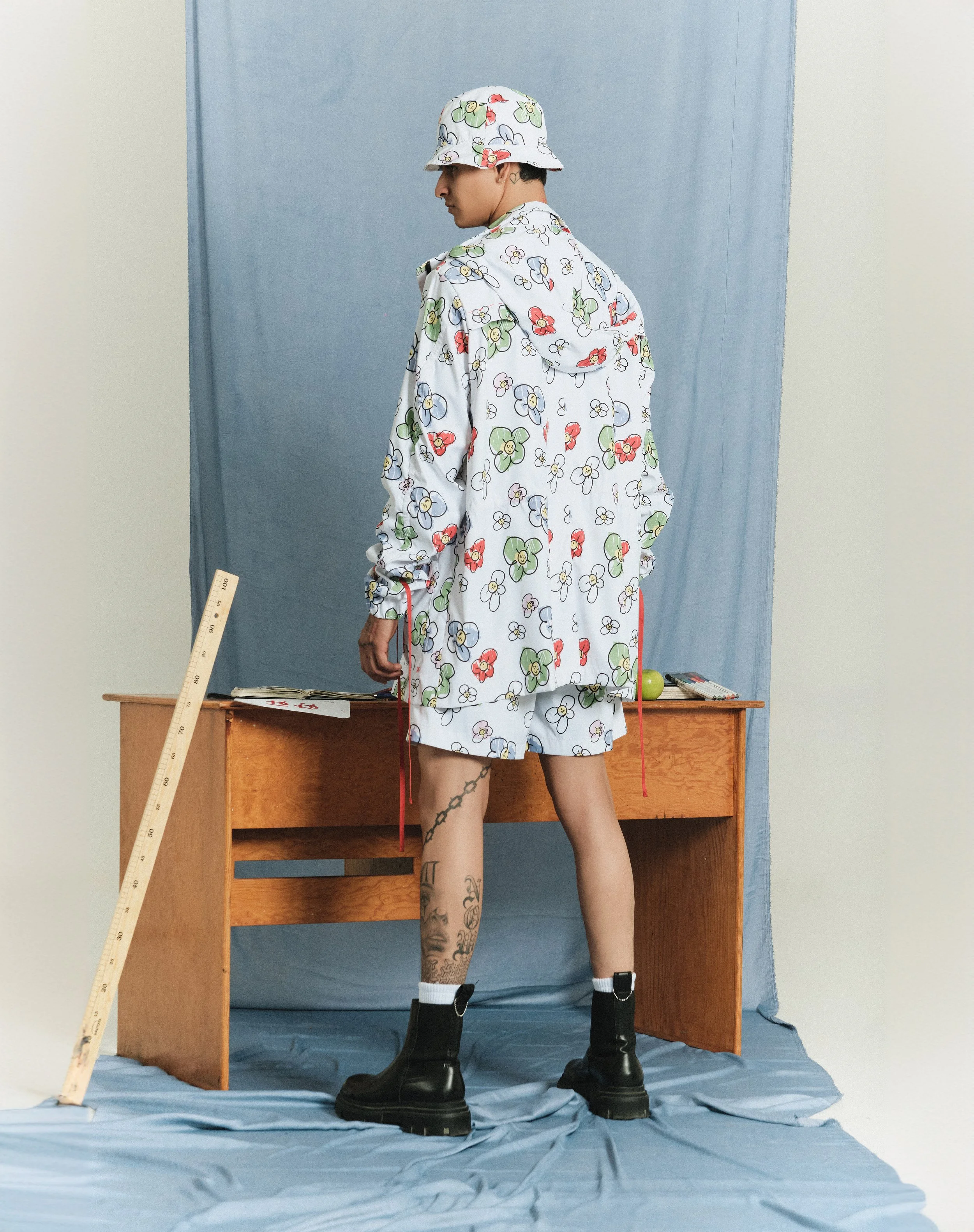 Una persona de espaldas lleva un conjunto de ropa con estampado floral, sombrero a juego, botas negras y calcetas blancas, de pie frente a un escritorio de madera con objetos sobre él, en un fondo de tela azul.