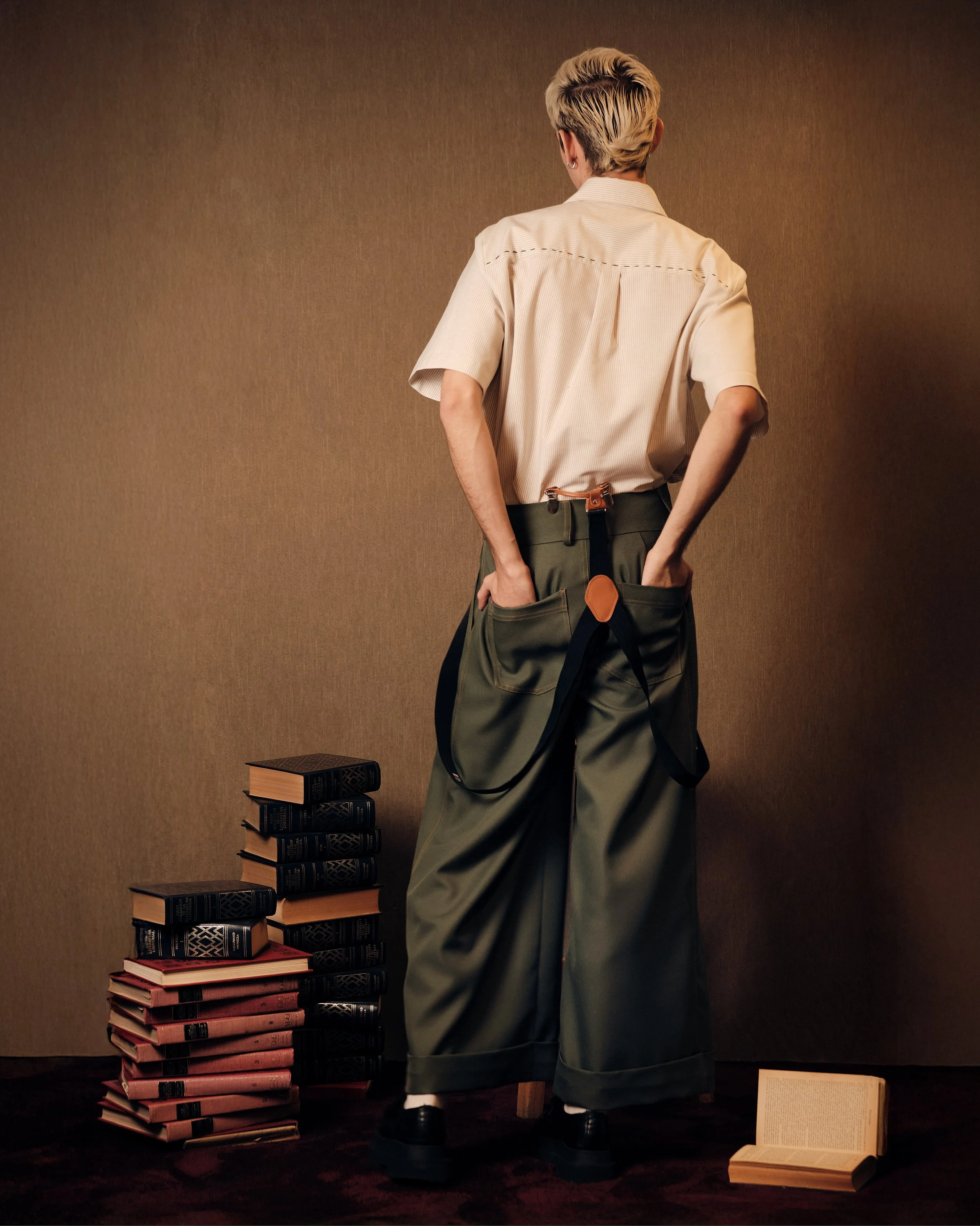 Persona de espaldas con cabello rubio platinado, pantalones anchos y cinturón con funda de cuero, rodeada de pilas de libros y un libro abierto en el suelo.