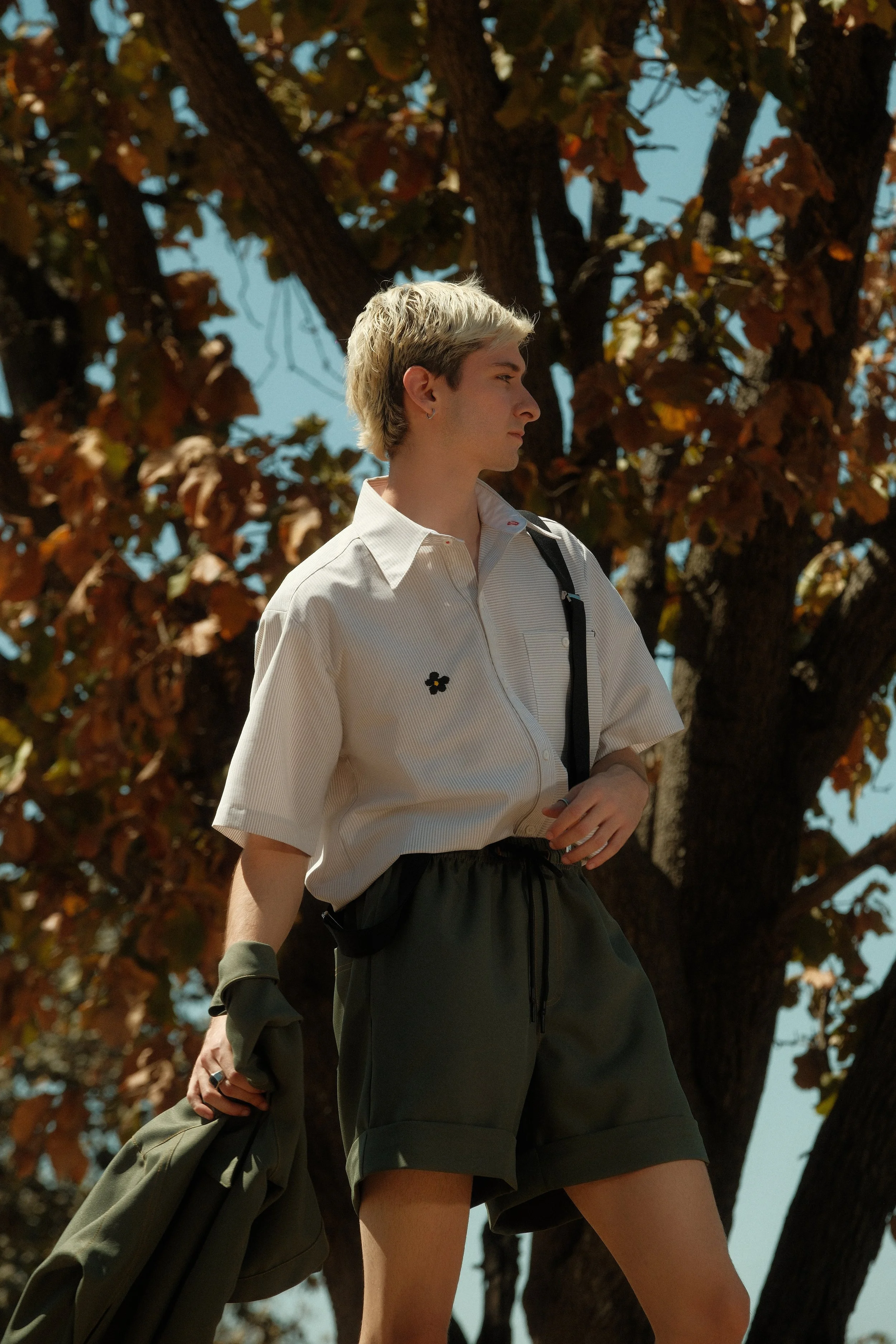 Joven con pelo rubio de espaldas, vistiendo blusa blanca y pantalones cortos, sosteniendo una prenda en la mano, de pie frente a un árbol con hojas secas.