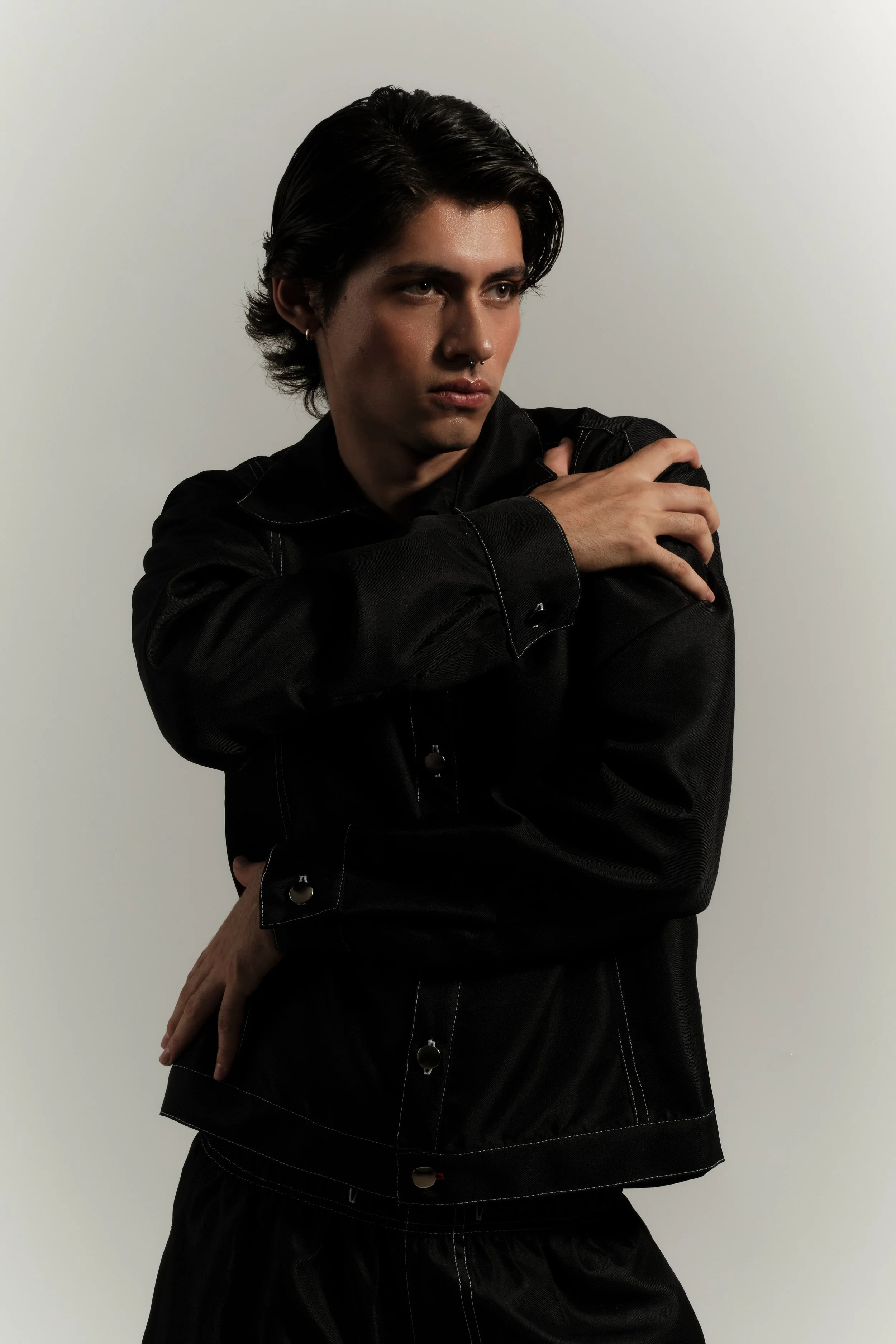 Joven con cabello oscuro y peinado hacia atrás, usando ropa negra, posando con una mano en el hombro opuesto, en un fondo gris claro