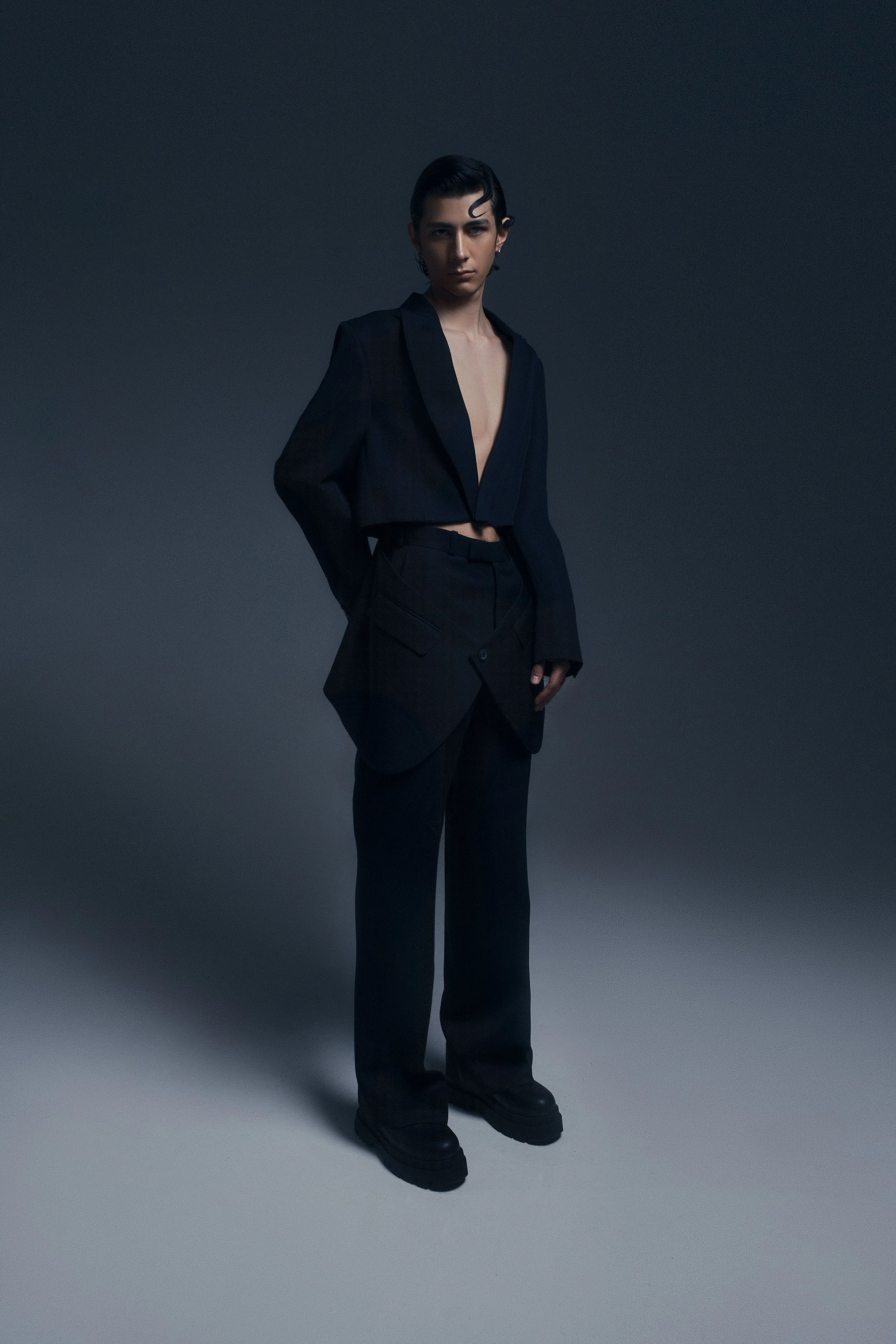 Persona con estilo andrógino usando un saco oscuro sin camiseta en un fondo gris oscuro.