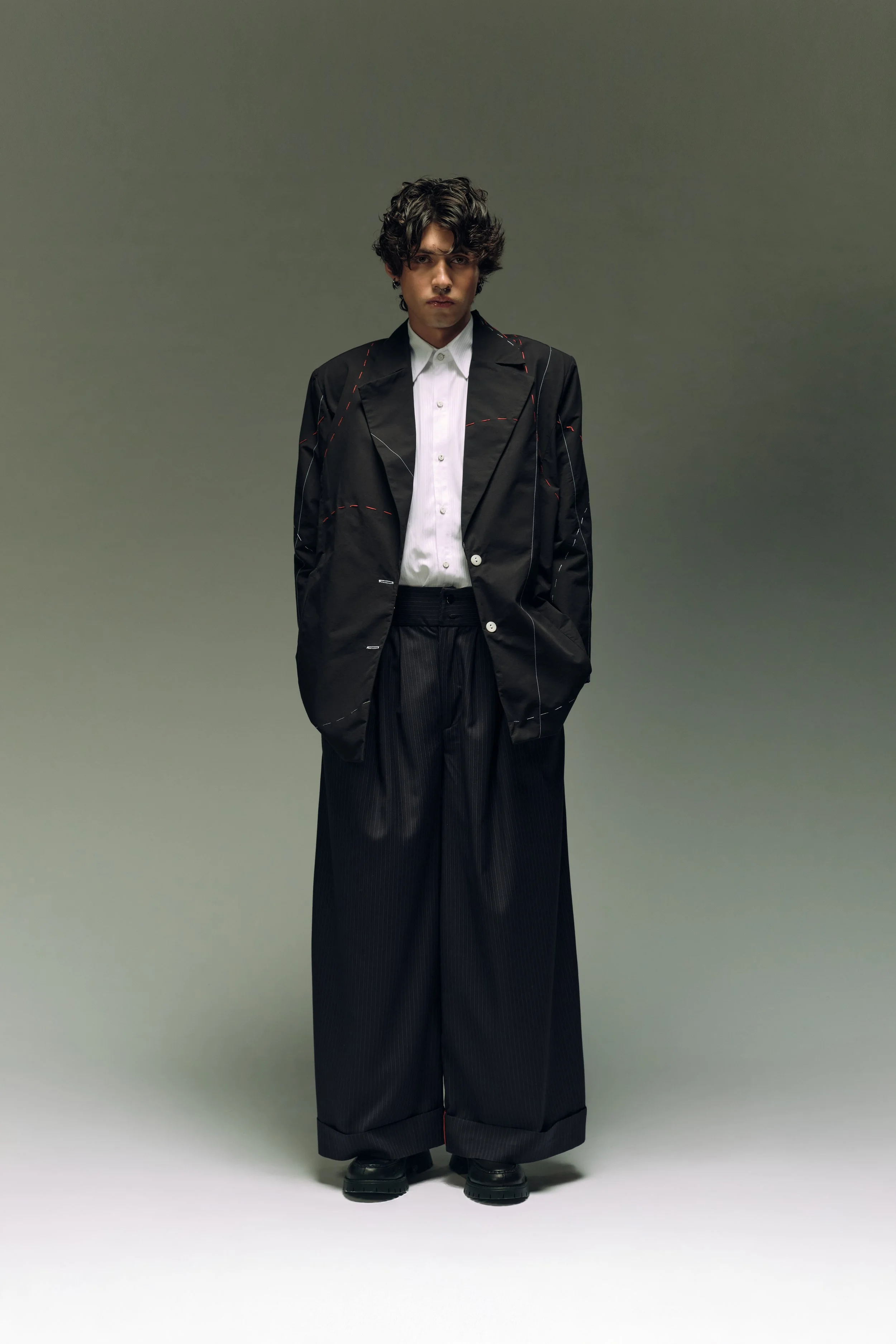 Hombre joven con cabello oscuro peinado al estilo desordenado, vestido con un saco negro con costuras y pantalones anchos negros, y una camisa blanca, de pie con las manos en los bolsillos, en un fondo gris.