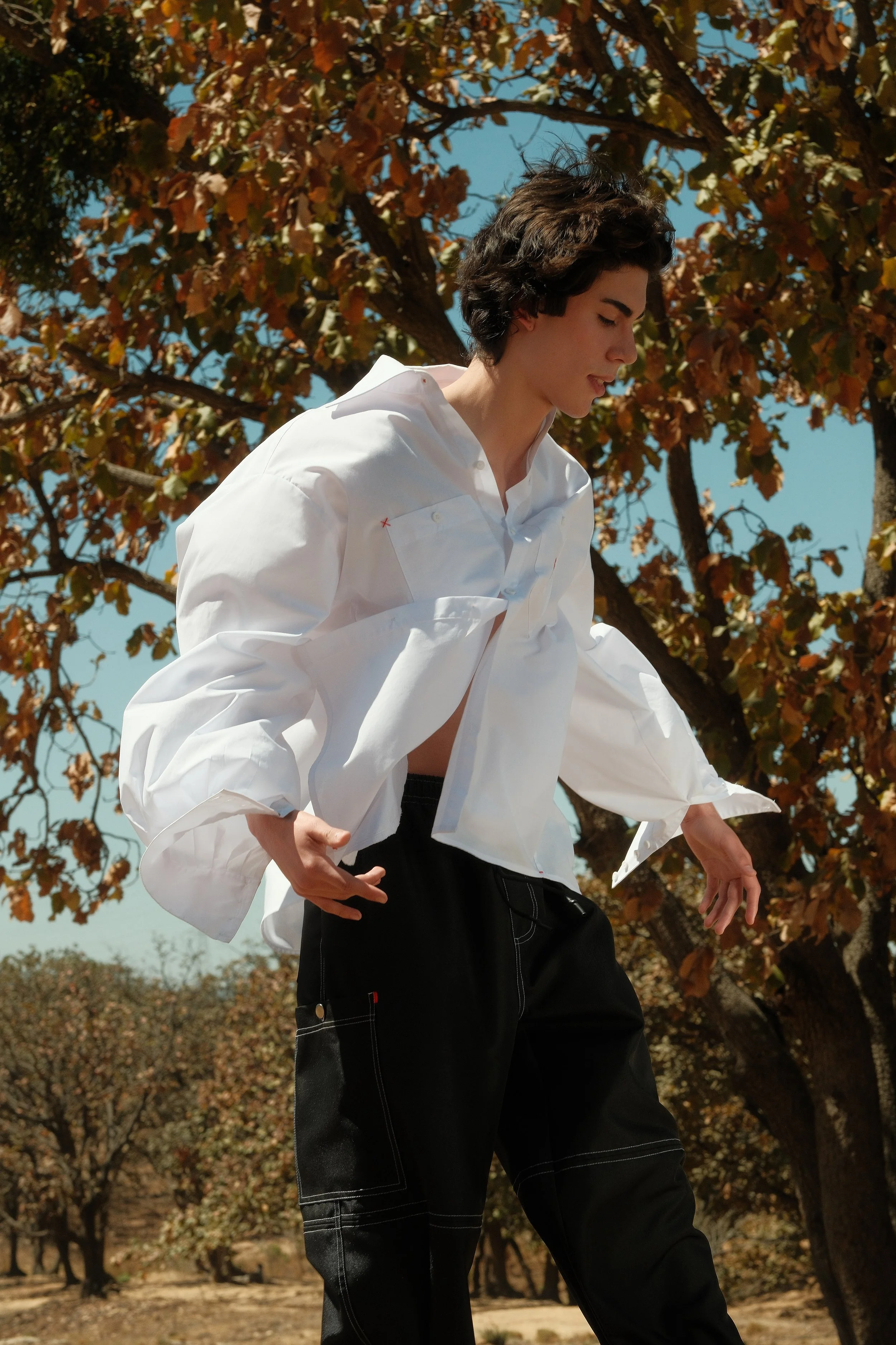 Persona joven con cabello corto, usando una blusa blanca y pantalones oscuros, de pie en un campo con árboles de hojas secas y cielo azul.