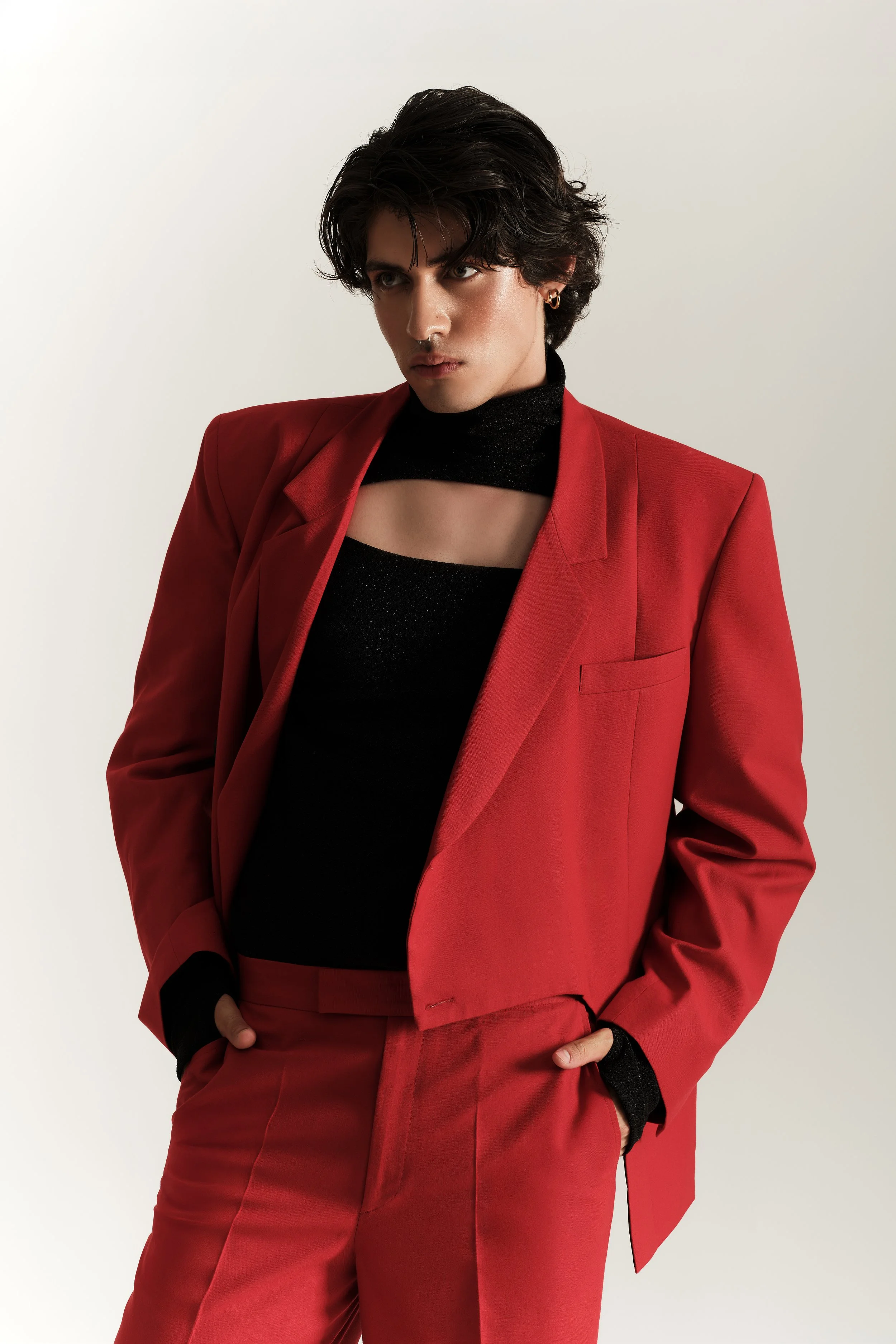 Un joven con cabello oscuro lleva un saco y pantalones rojos, y una camiseta negra con un recorte en el cuello, posando contra un fondo claro.