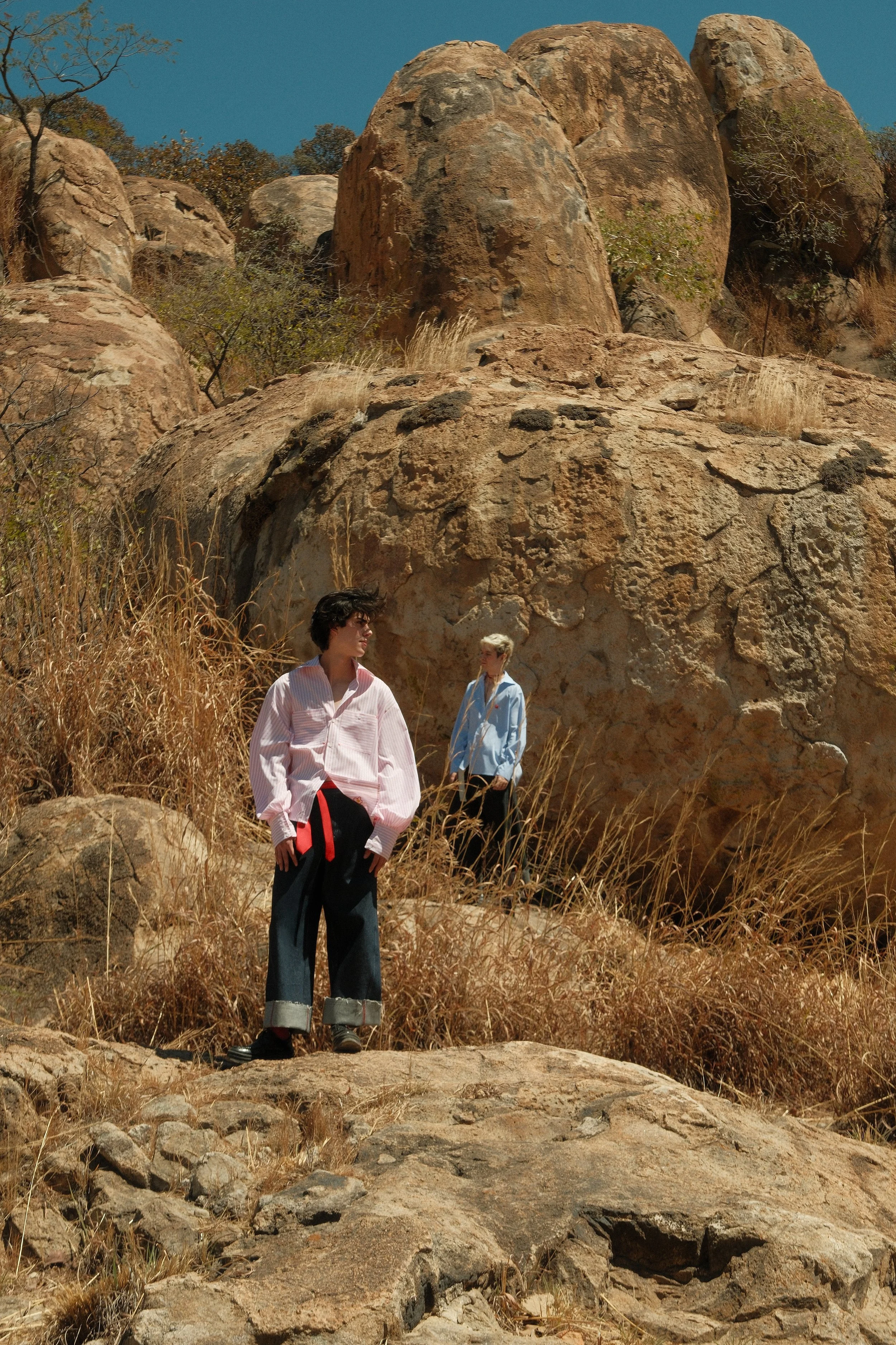 Dos mujeres de pie en un paisaje rocoso y árido con grandes rocas y vegetación seca bajo un cielo azul.