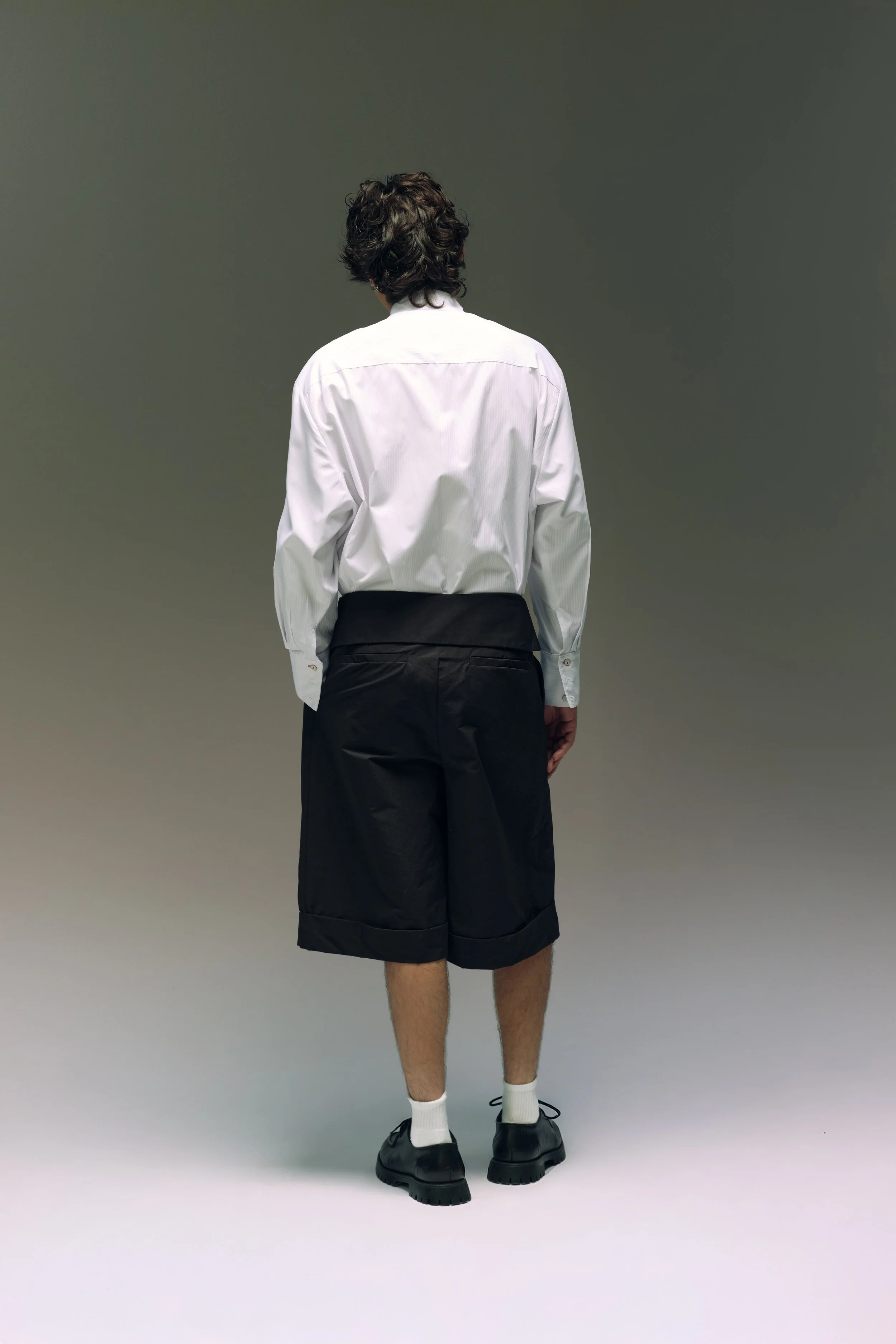 Persona de espaldas usando camisa blanca, pantalones cortos negros, calcetas blancas y zapatos negros en un fondo neutro.