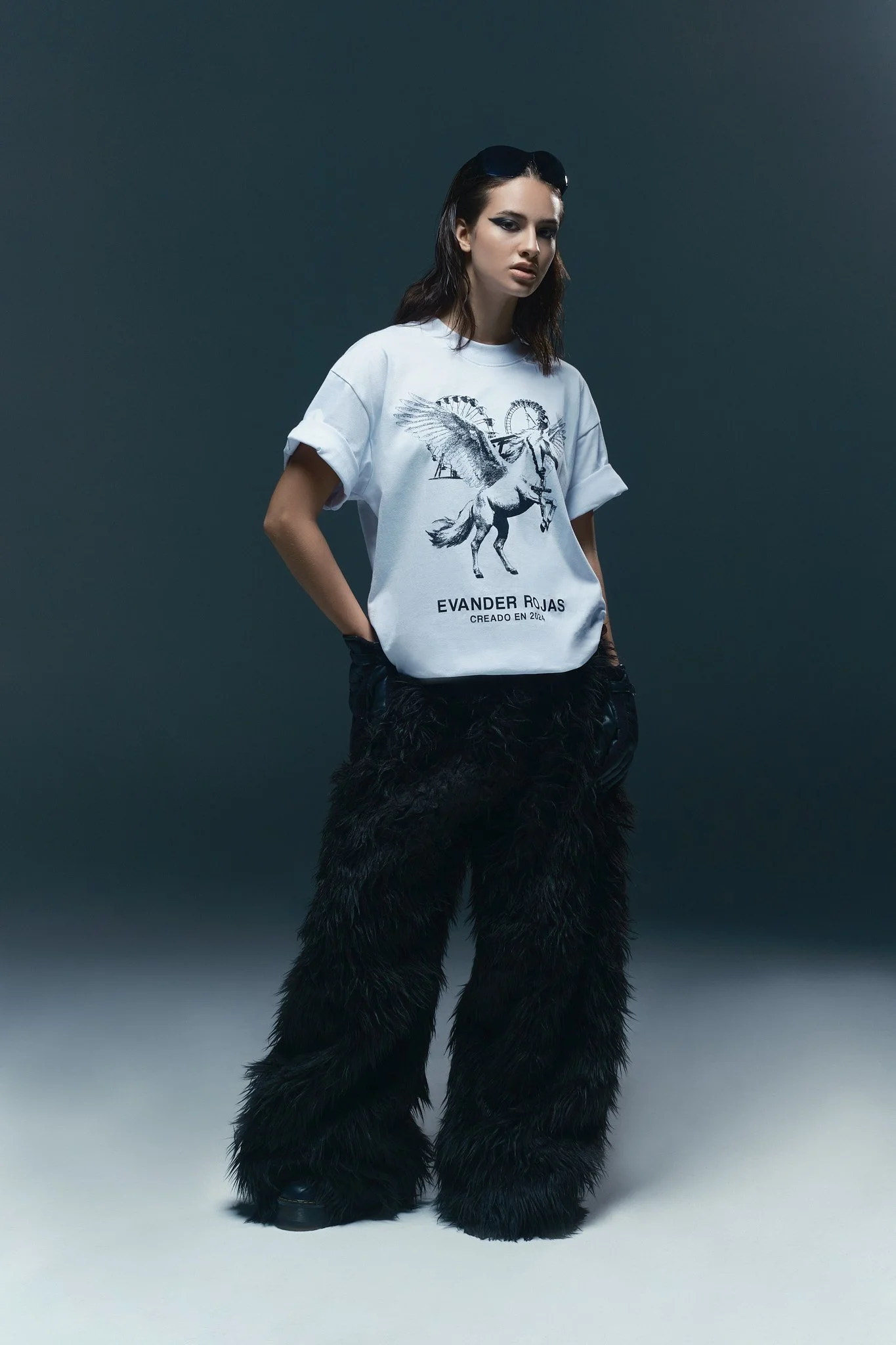 Mujer con gafas oscuras, camiseta blanca con diseño de unicornio y rueda de feria, pantalones de peluche negro y zapatos oscuros, posando de pie ante fondo oscuro.