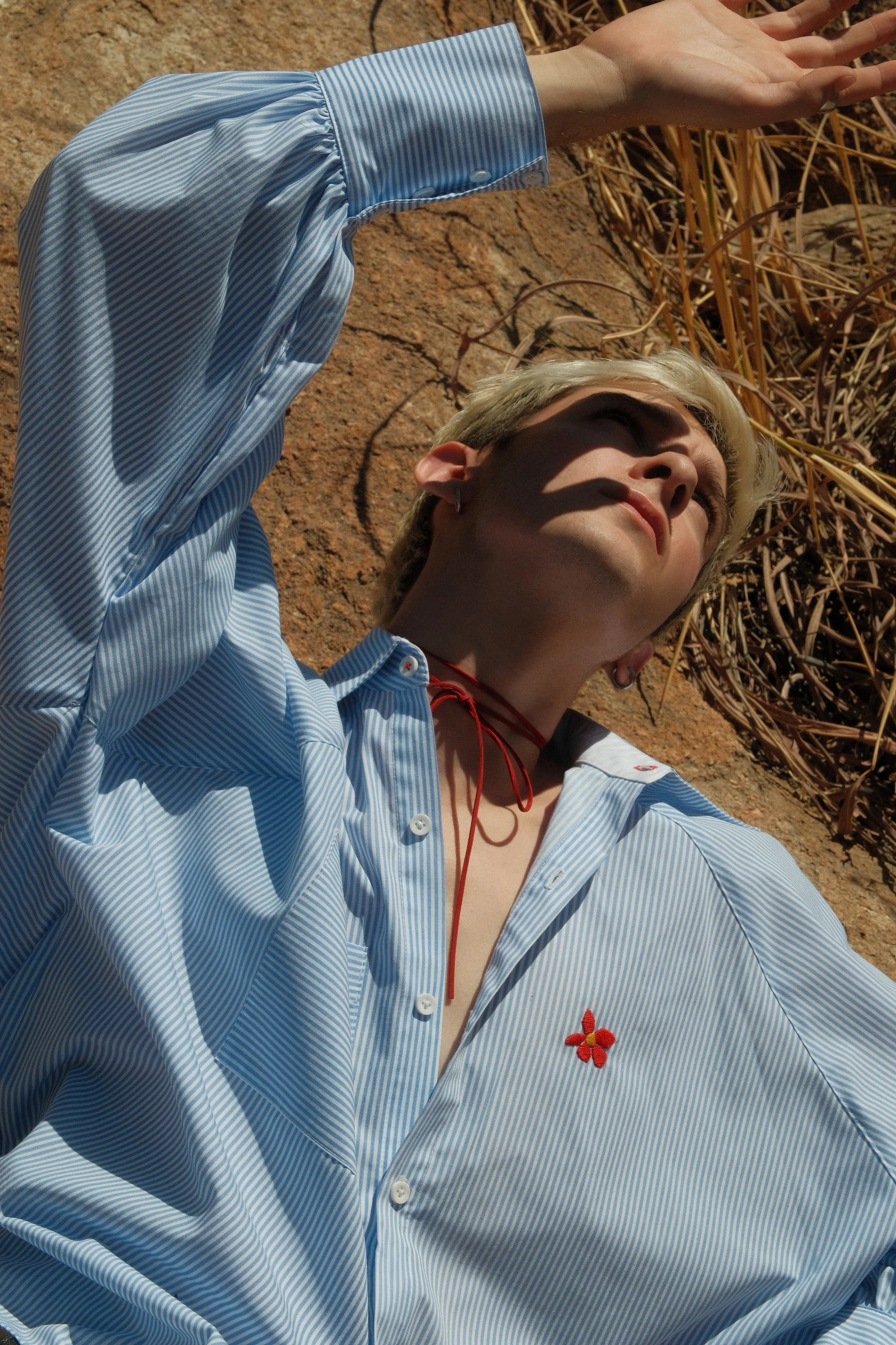 Persona con cabello rubio y camiseta de rayas azul y blanca, recostada en tierra arenosa, con sombra en su rostro, usando un collar rojo y pendientes.
