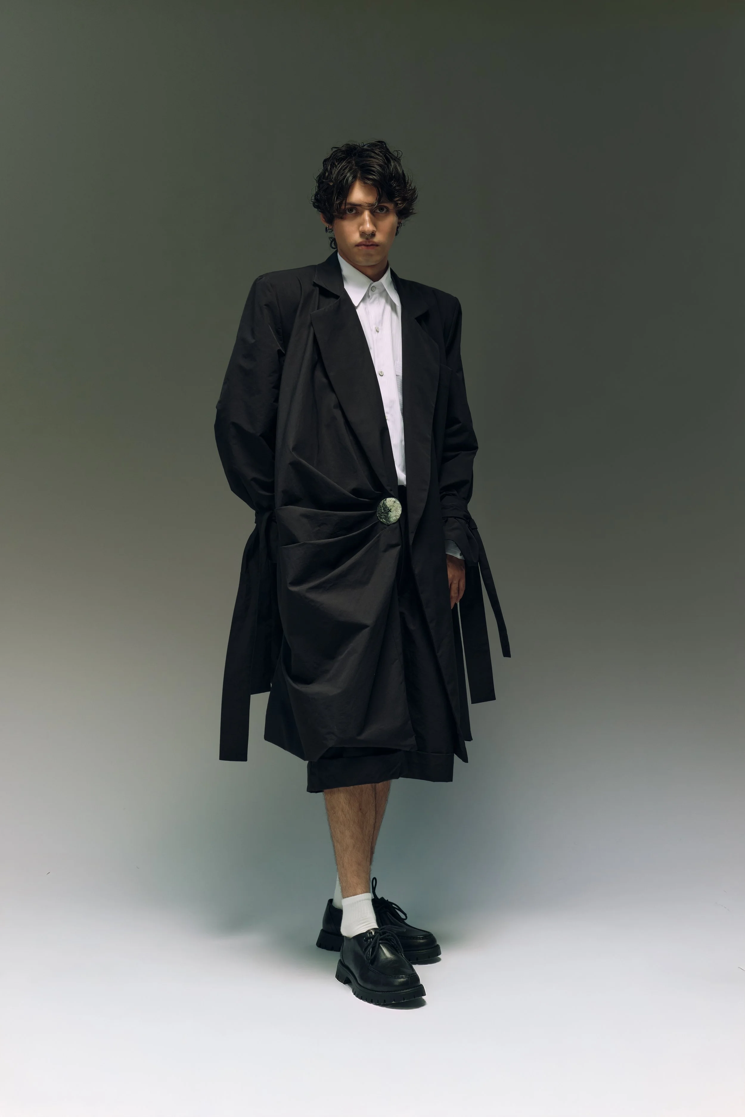 Joven vestido con ropa de moda, usa un abrigo largo negro y zapatos negros, en un fondo liso. 