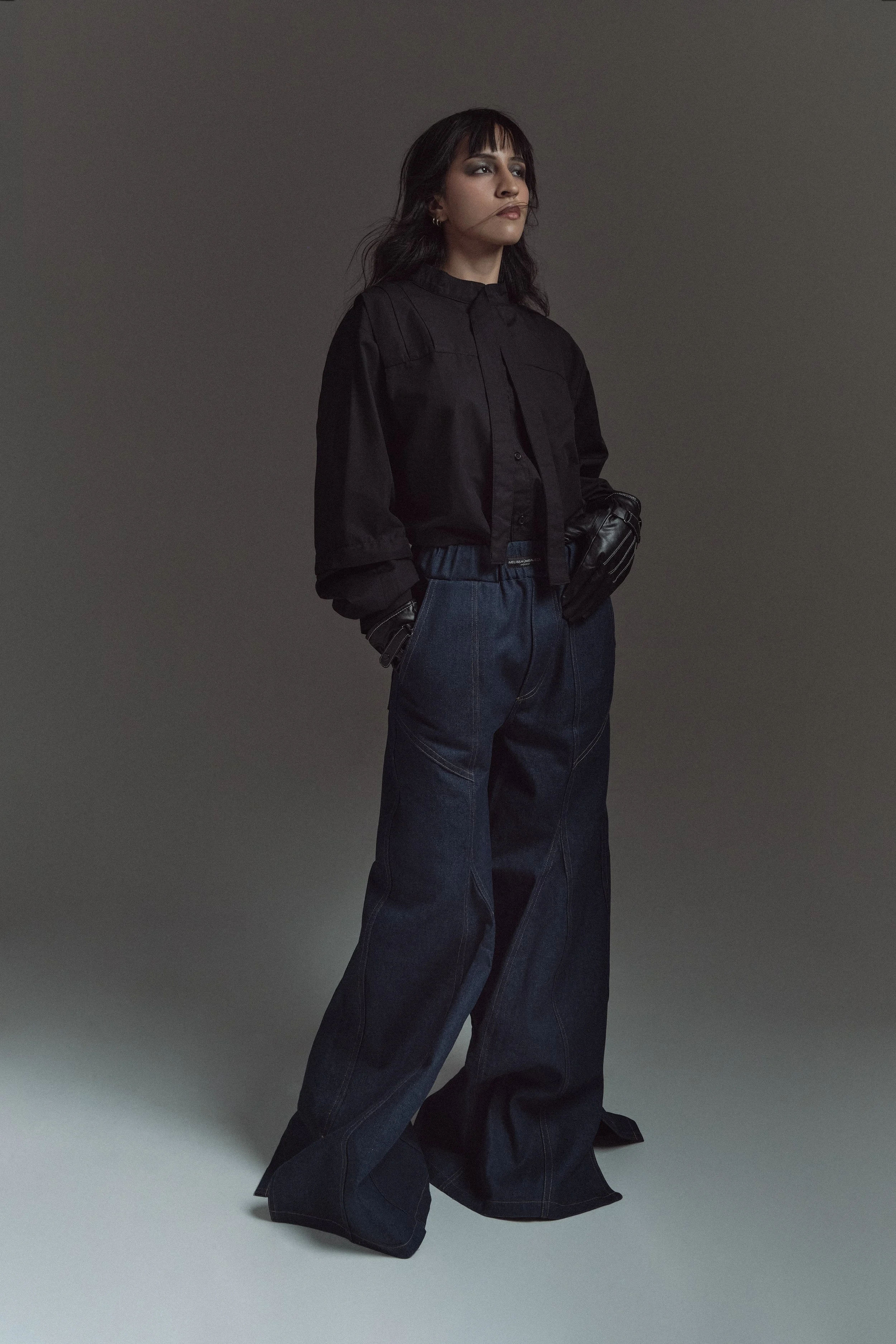 Mujer joven con cabello largo oscuro, vestida con una chaqueta negra y pantalones anchos, en un fondo gris oscuro.