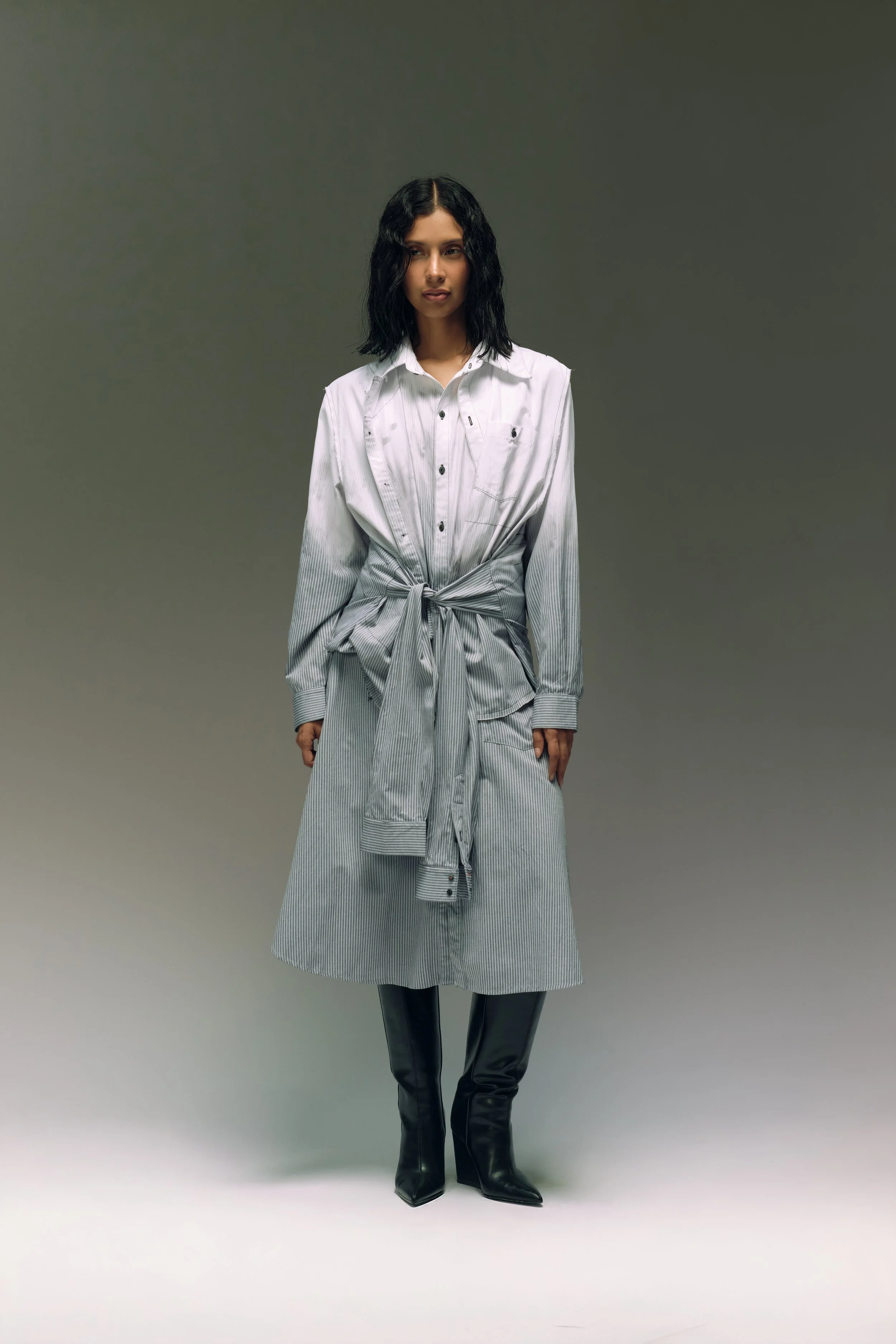 Mujer con vestido y botas negras, parada en un fondo gris.