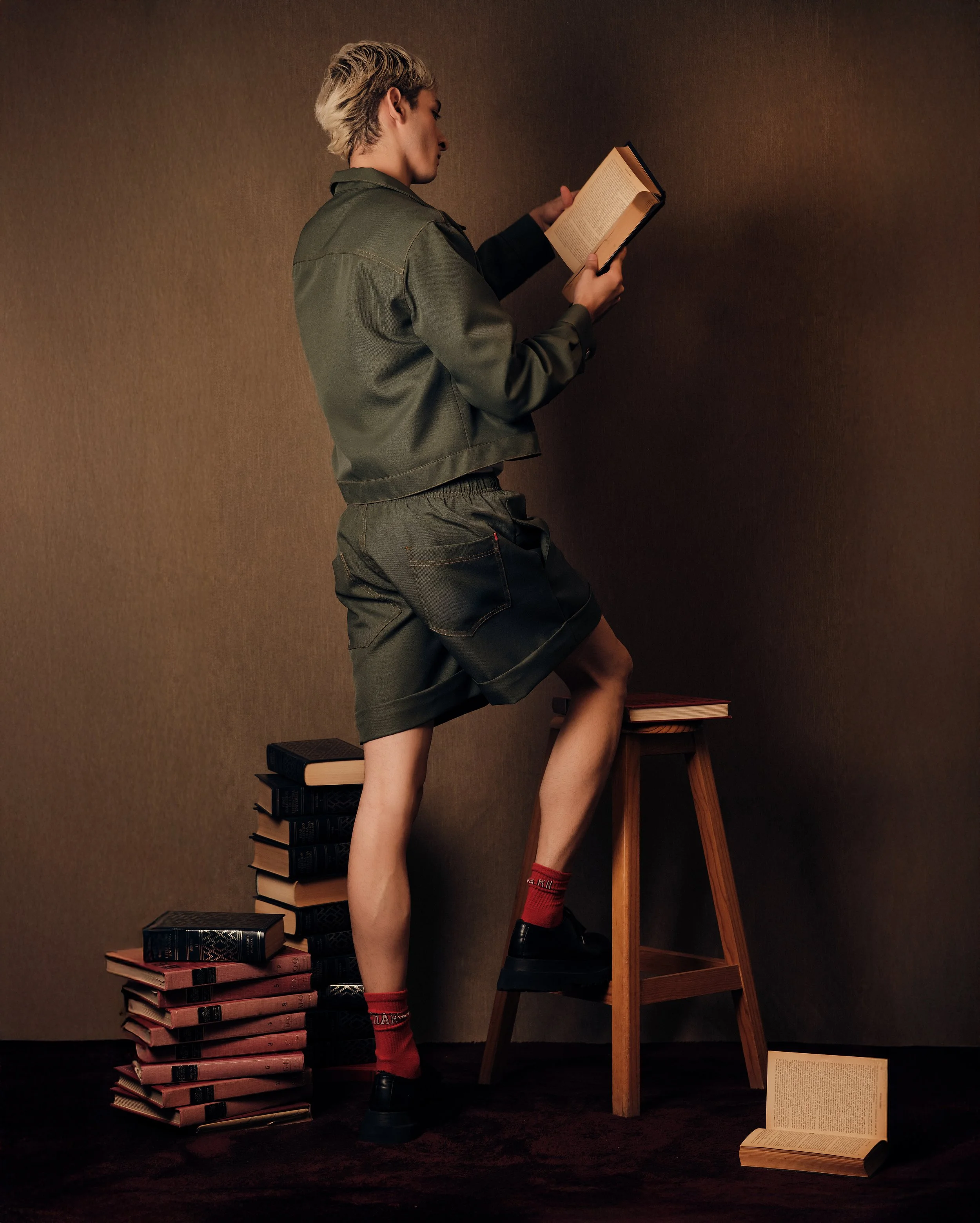 Persona leyendo un libro en un entorno con muchas pilas de libros, con un fondo de pared marrón.