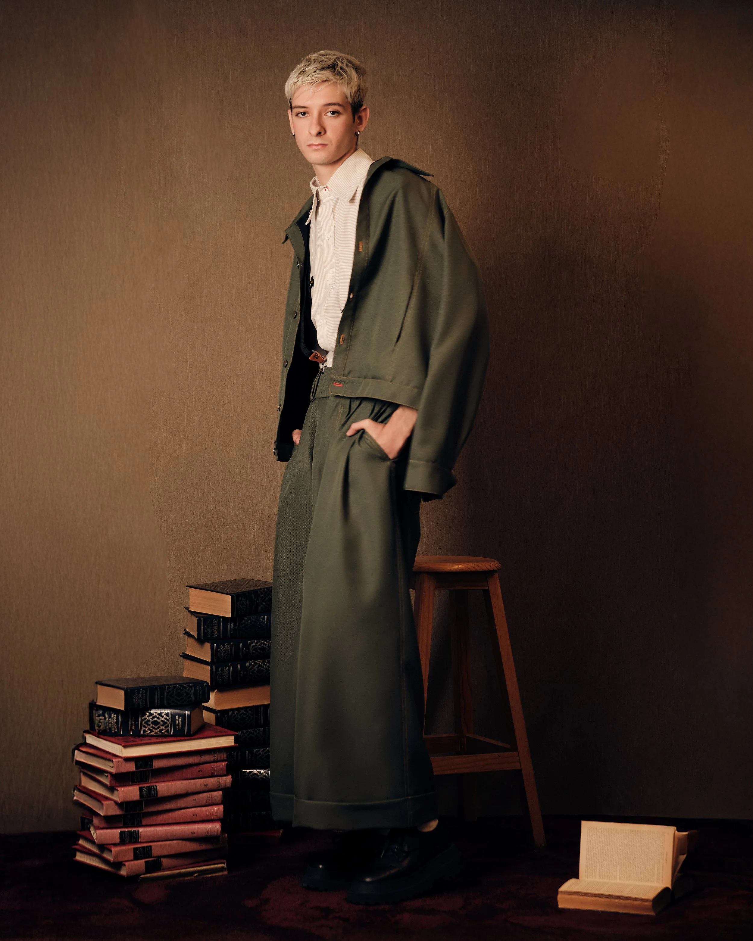 Una persona con cabello rubio y corto, vestida con pantalones anchos y chaqueta verde, de pie junto a una pila de libros y un taburete de madera en una habitación con paredes marrones.