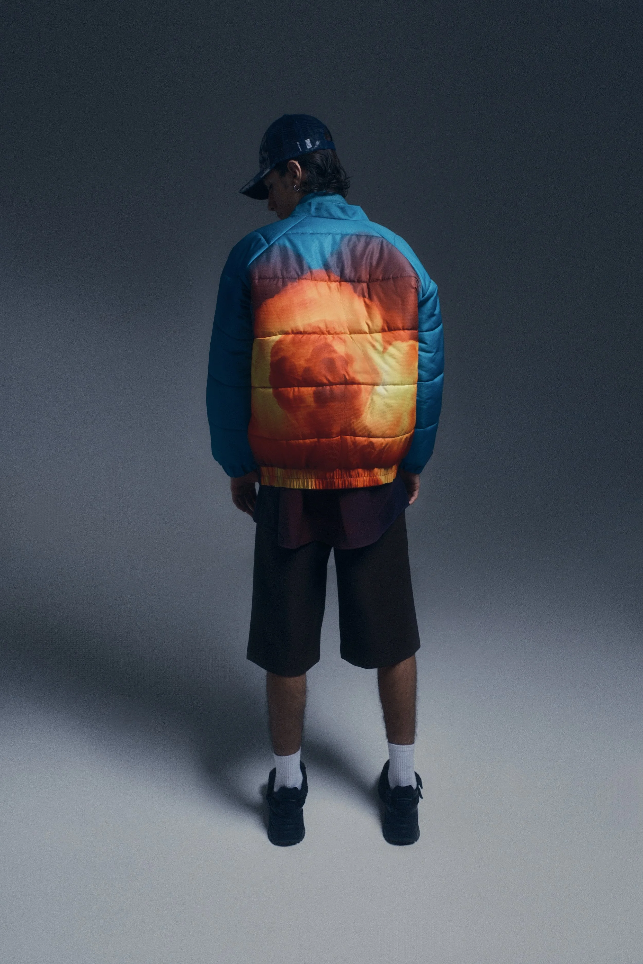 Persona de espaldas vistiendo una chaqueta azul con un estampado de un rostro en tonos naranjas y amarillos, pantalones cortos negros, calcetines blancos y zapatillas negras, en un fondo gris oscuro.