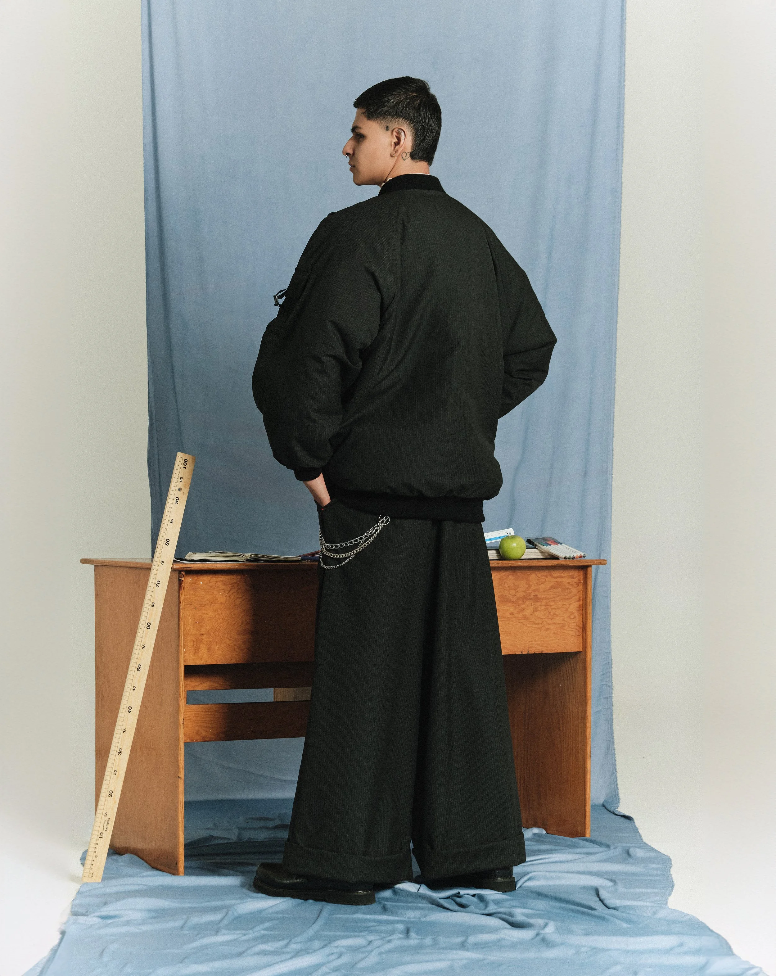 Persona de espaldas con ropa negra en un entorno con fondo azul y una mesa de madera con objetos como libros, un cuaderno, y una manzana