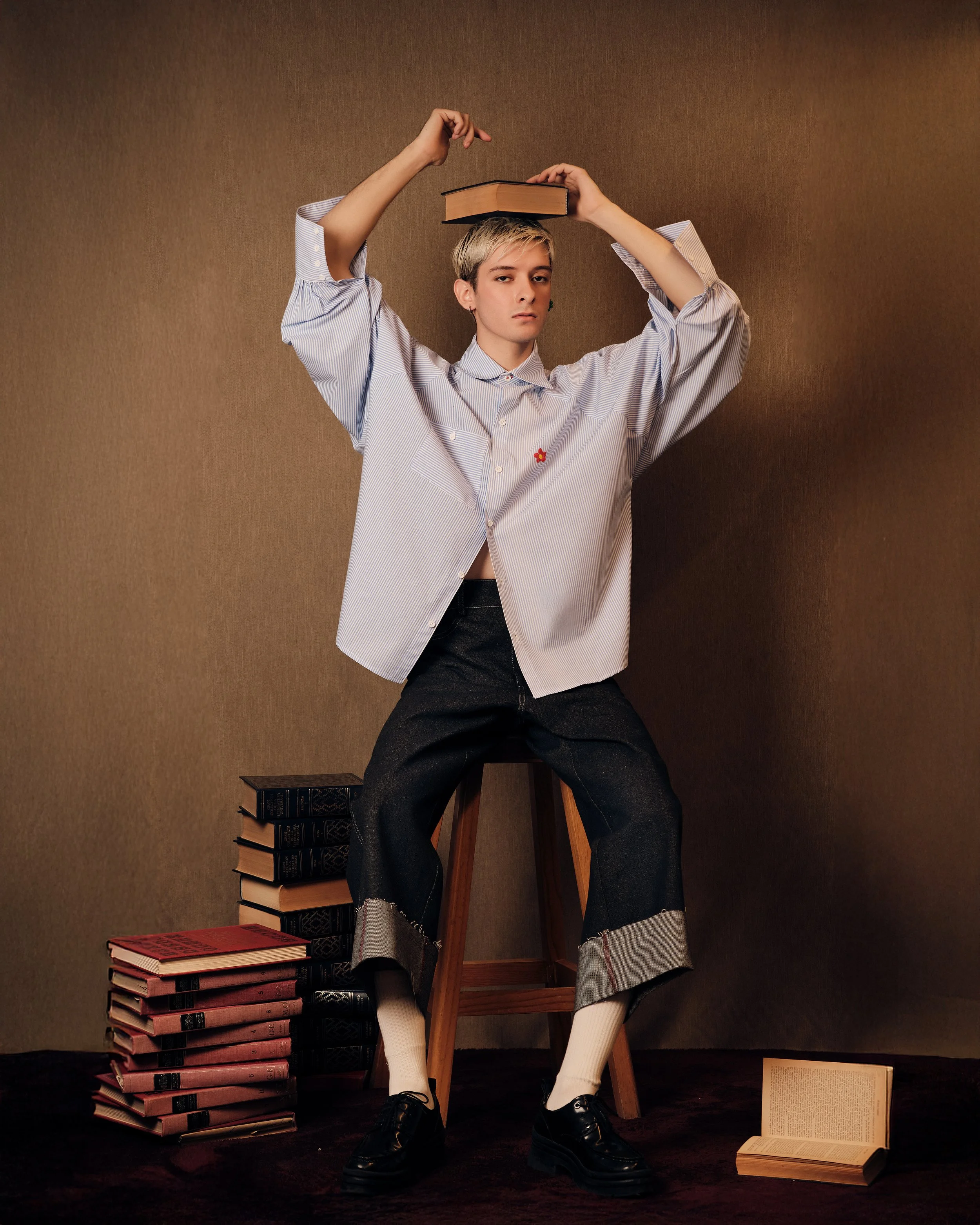 Joven sentado en un banquito con libros apilados a su lado y uno abierto en el suelo, usando una camisa y pantalones cortos, con un libro en la cabeza, en una pose artística.