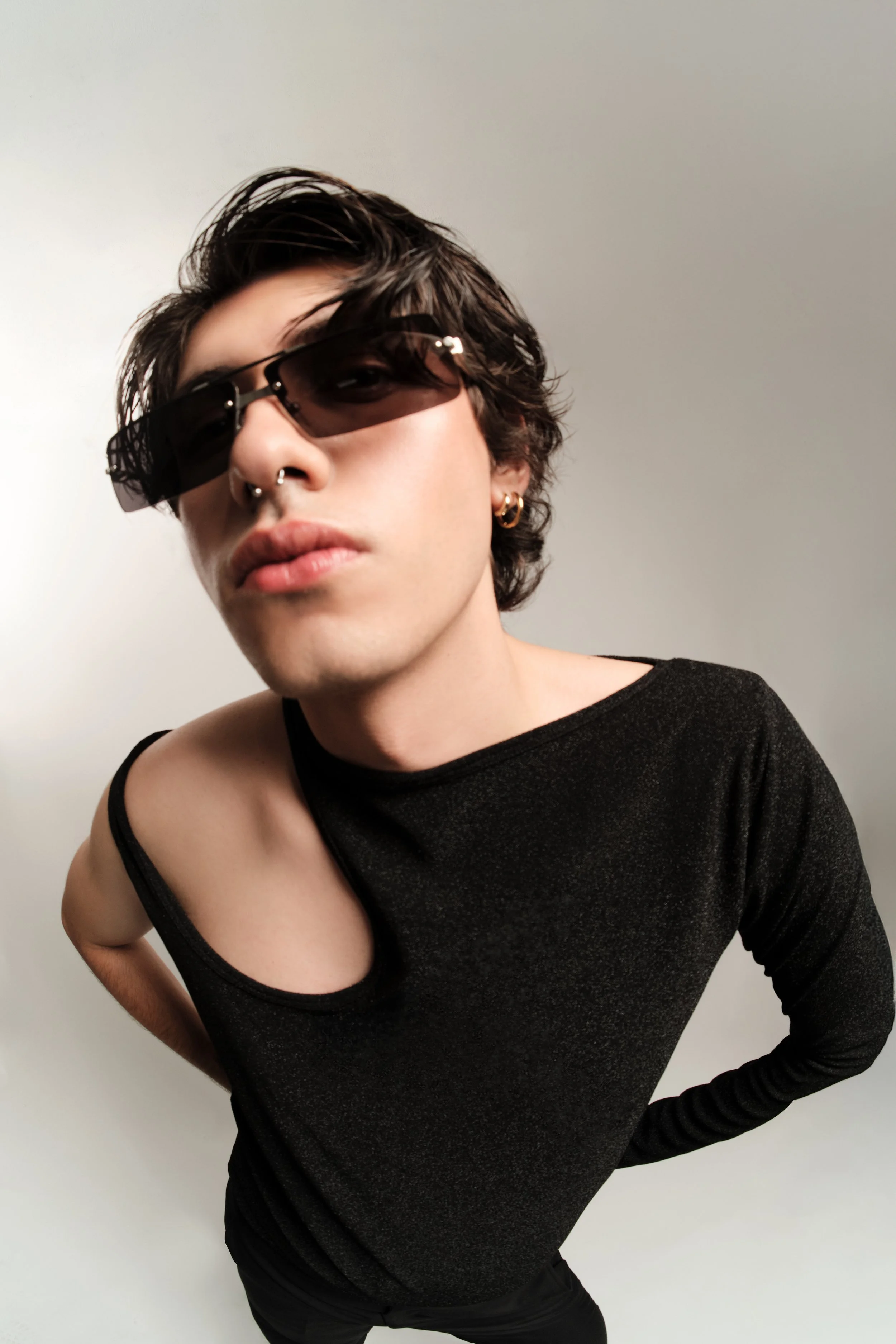 Joven con gafas de sol oscuras, peinado desordenado, piercings y ropa negra, en posición inclinada hacia adelante.