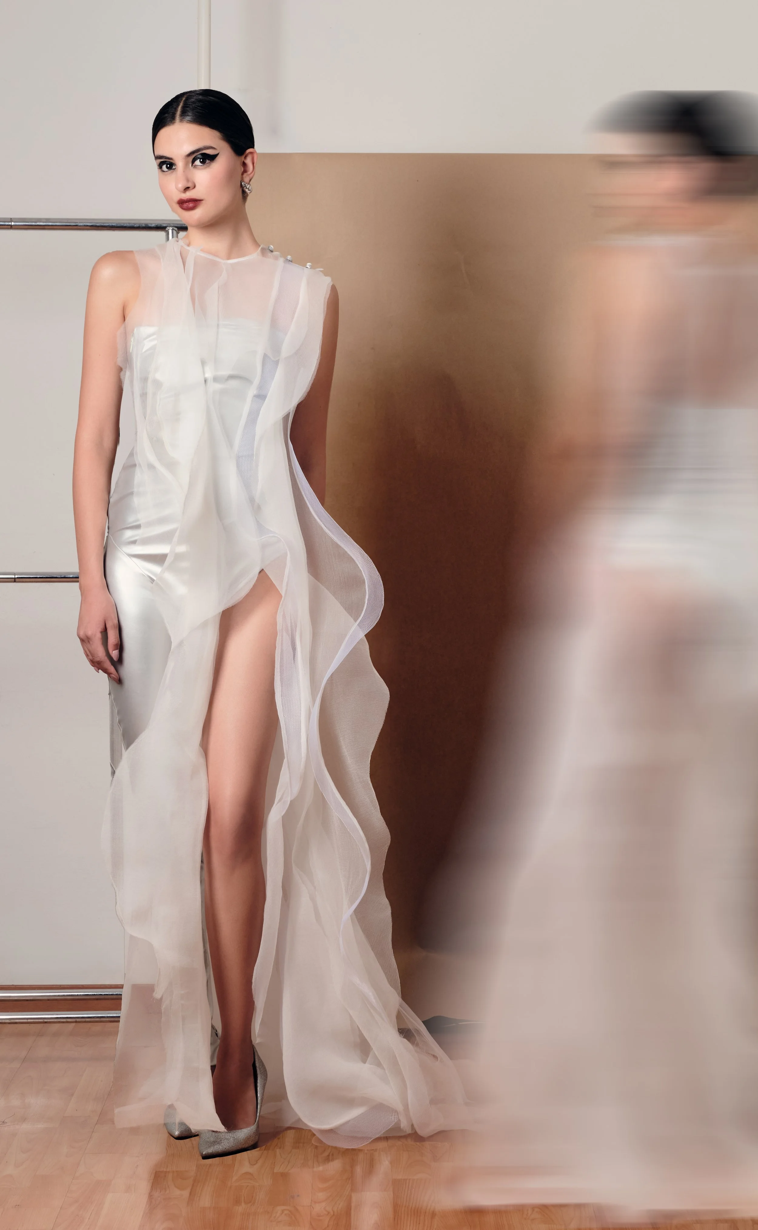 Mujer con vestido de noche de tela ligera y transparente, con un solo zapato plateado, posando en interior con pared beige y suelo de madera.