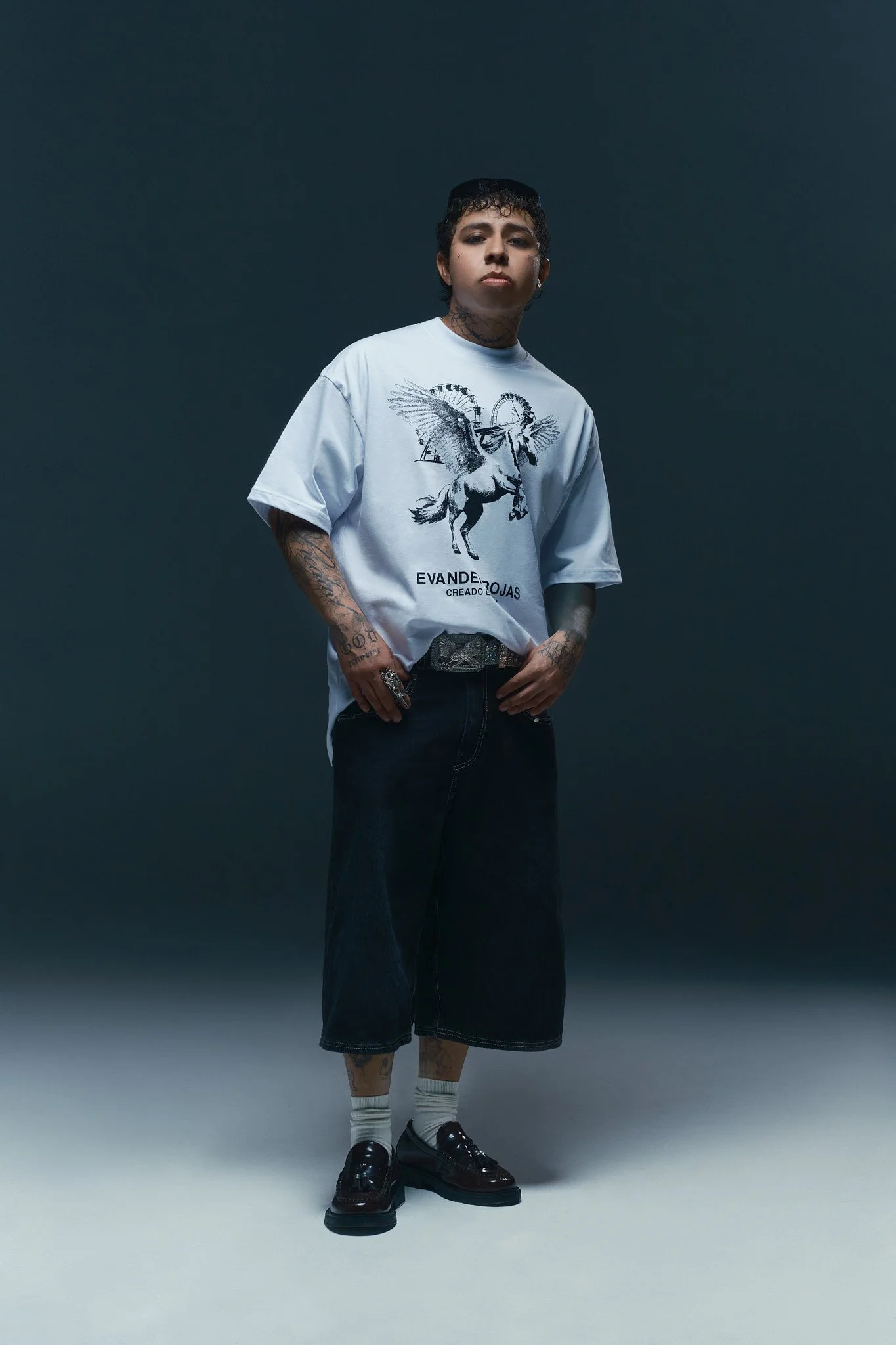 Joven con pantalones cortos negros, camiseta blanca con diseño de un caballo con alas y la palabra 'EVANDER' en letras grandes, zapatos negros brillantes, calcetas blancas, y pelo rizado corto, en fondo oscuro.