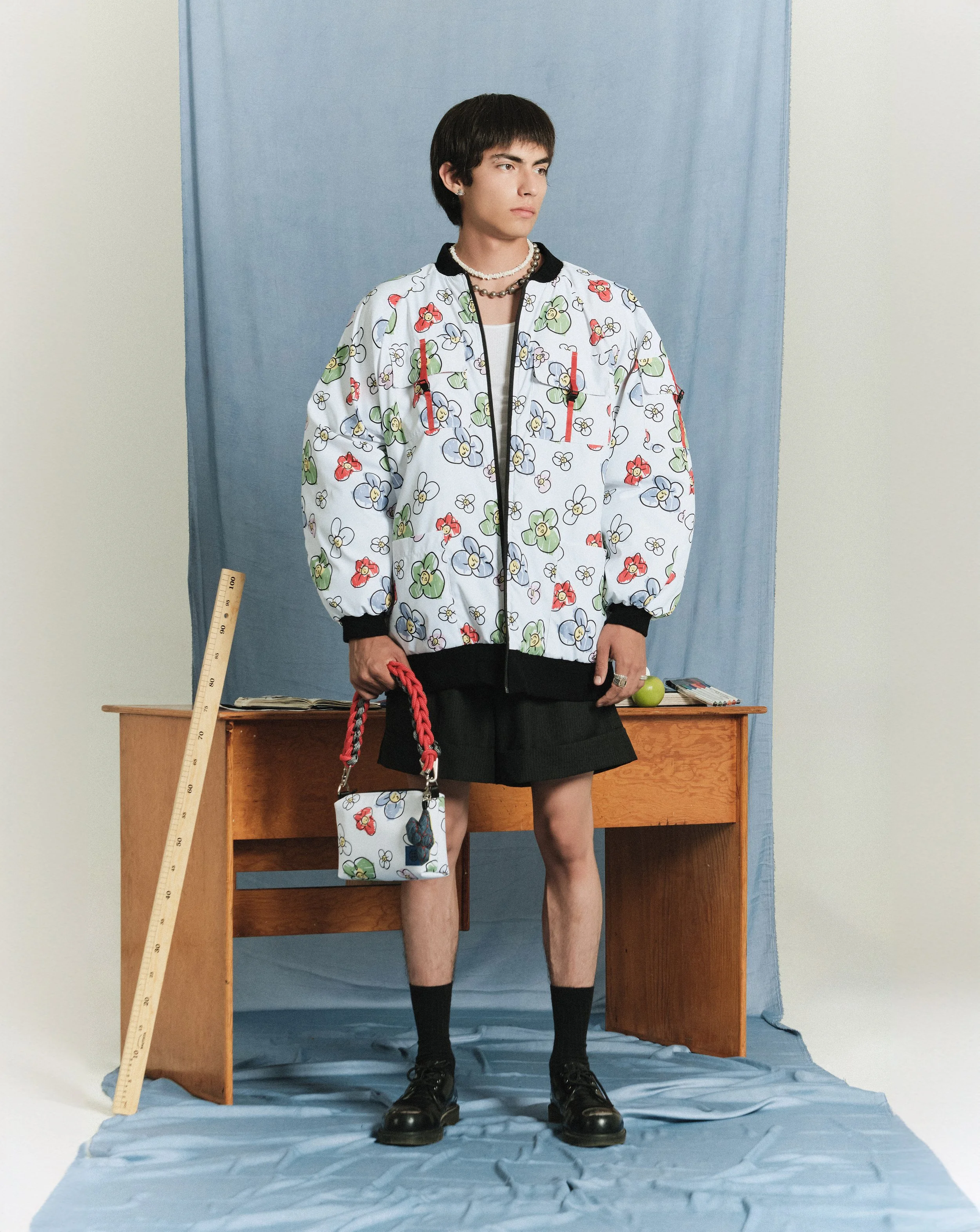 Joven con ropa de moda, chaqueta con estampado de flores, pantalón corto y accesorios en un entorno de estudio con fondo azul y objetos como regla y libro.