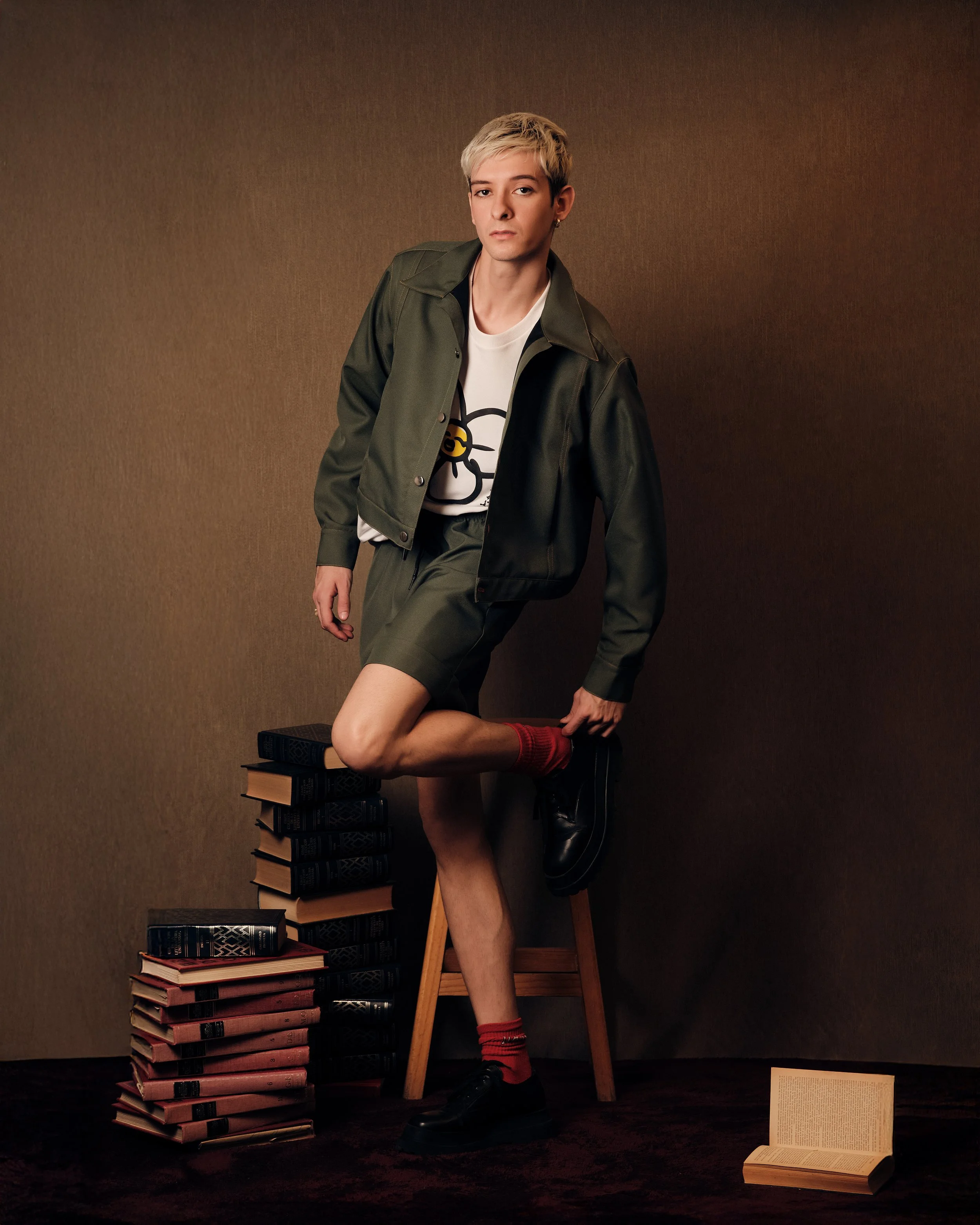 Joven con pelo rubio y peinado asimétrico, usando chaqueta verde, camiseta blanca con dibujo, pantalones cortos, calcetas rojas y zapatos negros, apoyado en una silla, rodeado de pilas de libros y un libro abierto en el suelo.