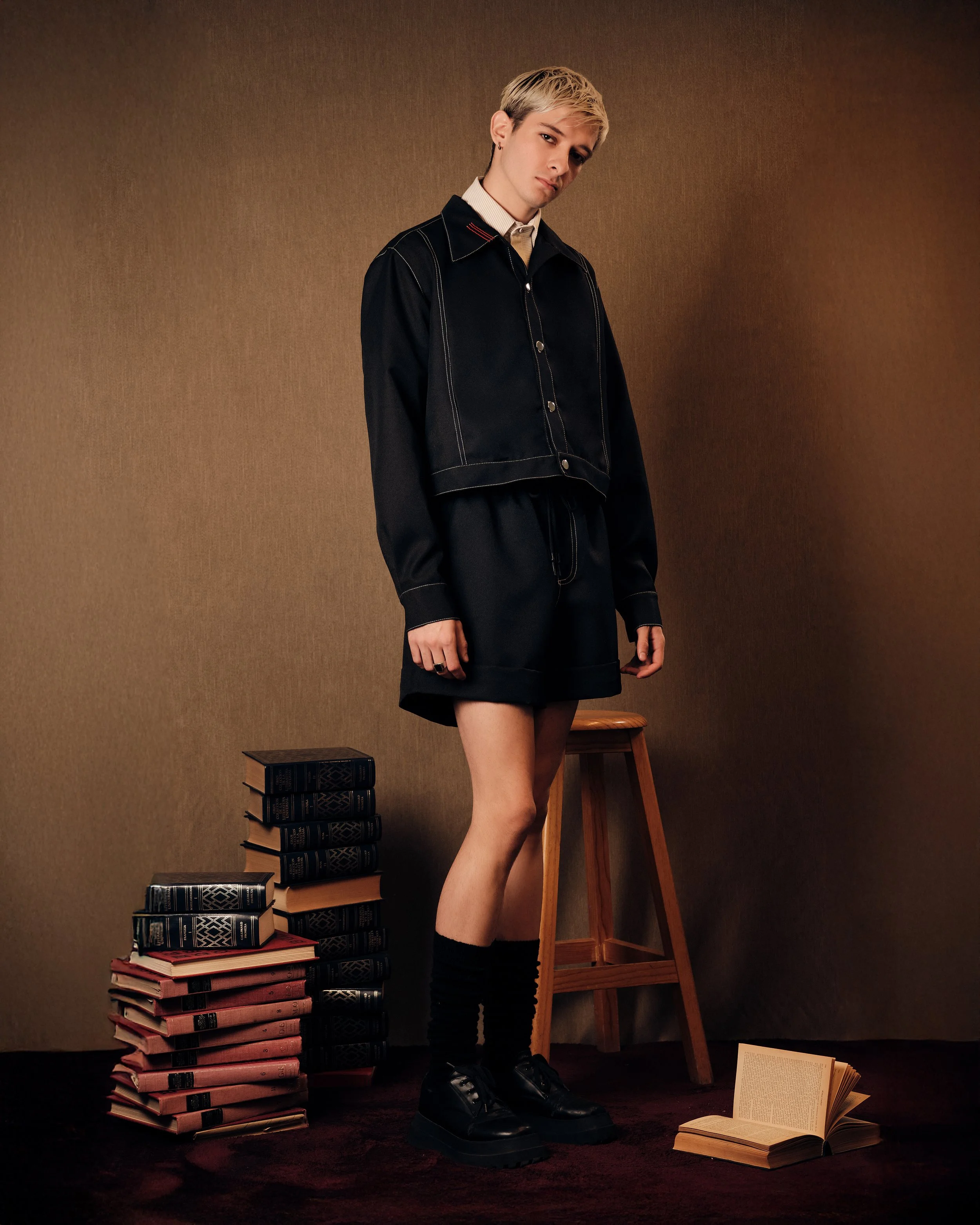 Persona de género masculino con cabello rubio y peinado a la moda, viste chaqueta negra, shorts cortos y botas negras, rodeada de pilas de libros y un libro abierto en el suelo en una habitación con pared de tono beige.