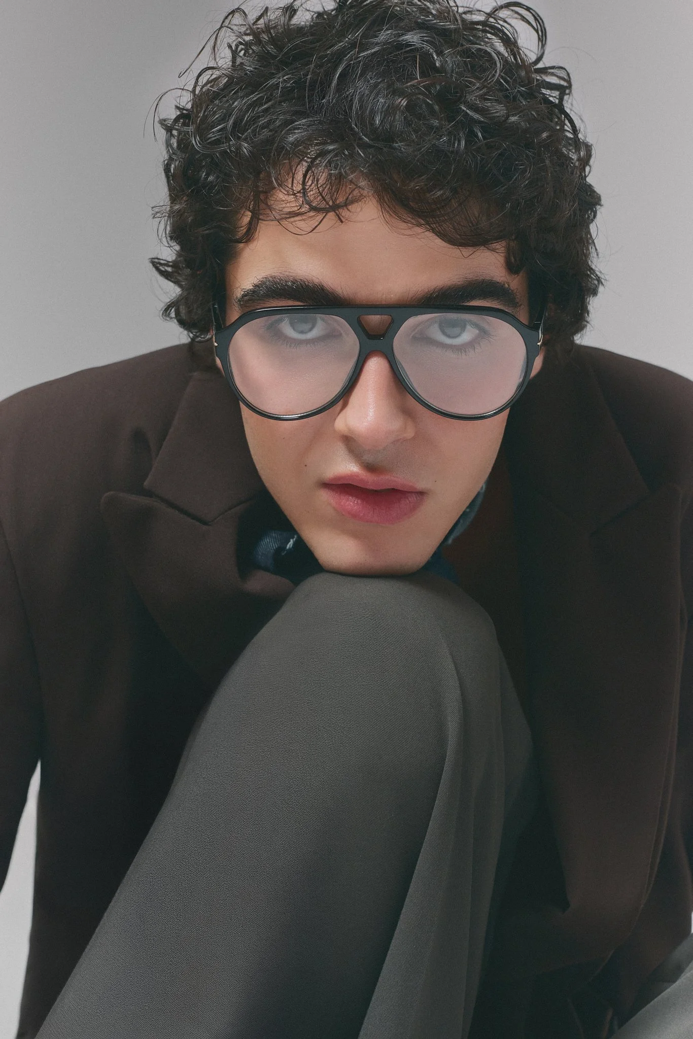 Retrato de un joven con cabello rizado, gafas grandes y expresión intensa, vestido con saco oscuro y pantalón gris