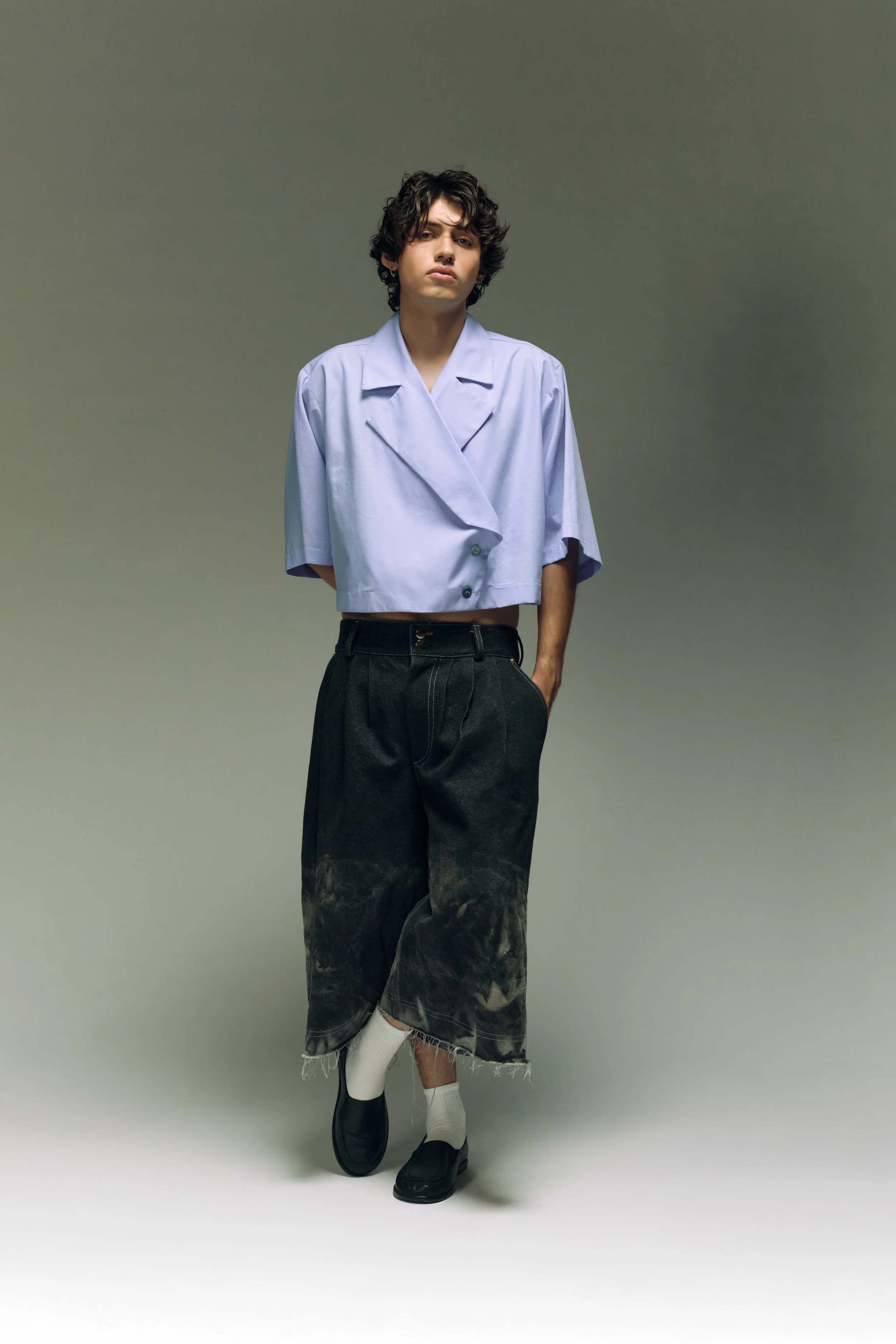 Persona joven con peinado rizado, usando blusa azul, pantalones anchos rotos, calcetas blancas y zapatos negros, posando en un fondo gris liso.
