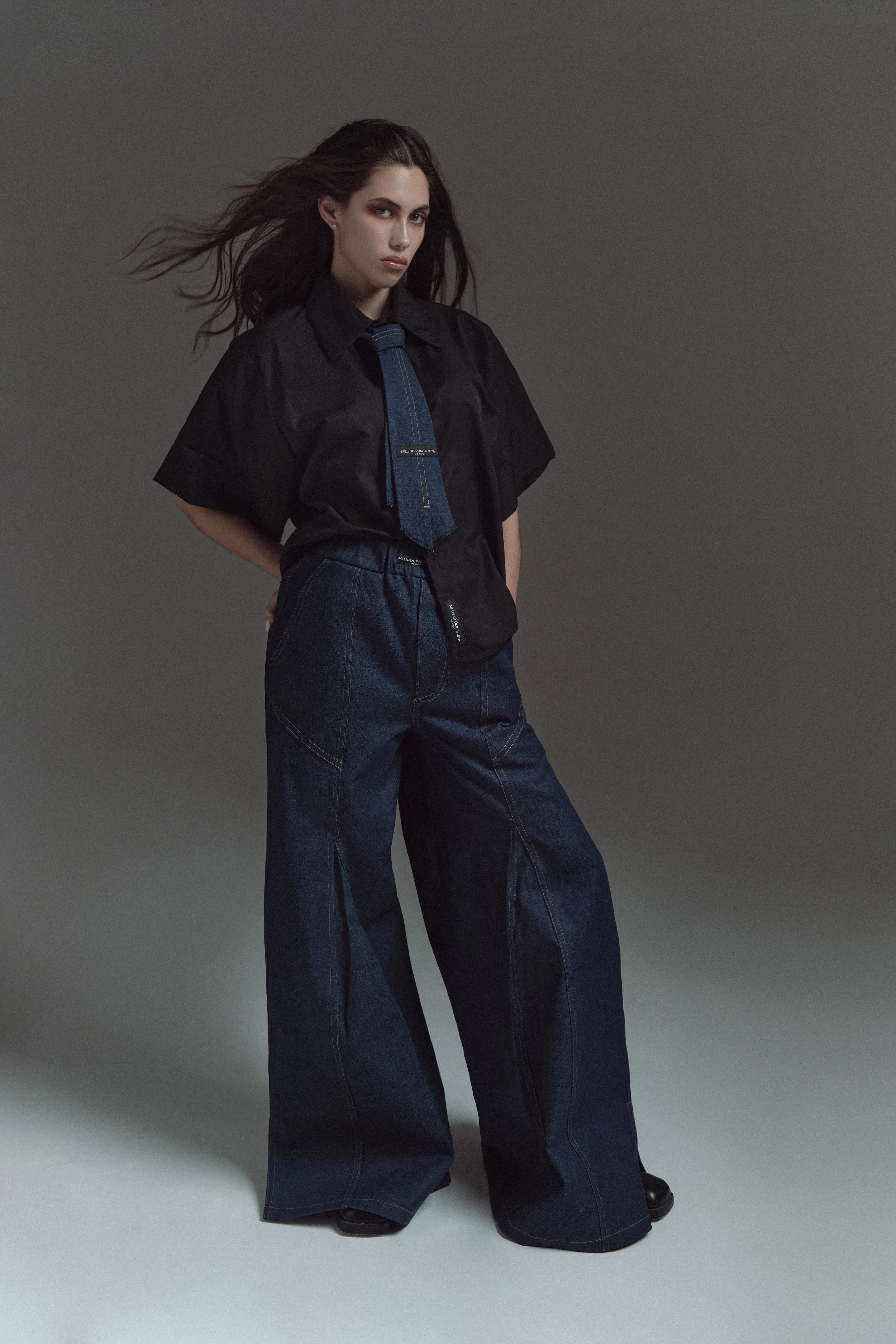 Retrato de una mujer con cabello largo y suelto, vestida con una camisa negra y pantalones de mezclilla anchos, en un fondo gris oscuro.