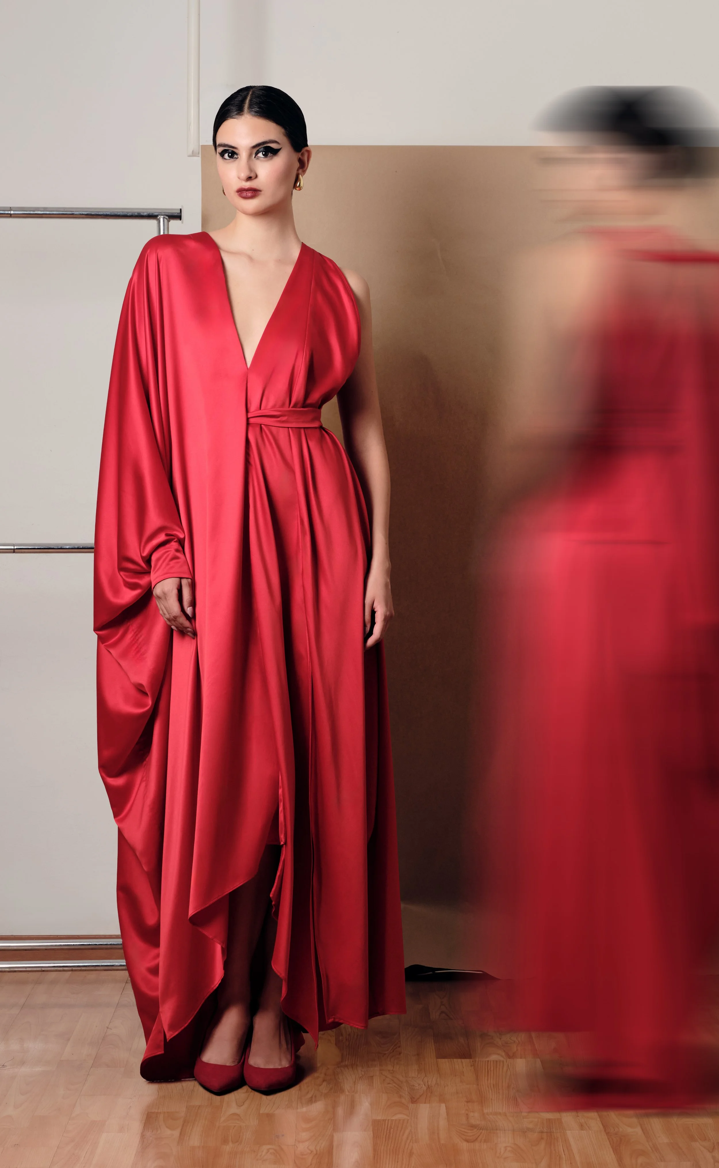 Mujer con vestido largo de satén rojo y tacones del mismo color, con cabello peinado hacia atrás, en un ambiente interior, con una pared beige y un suelo de madera.