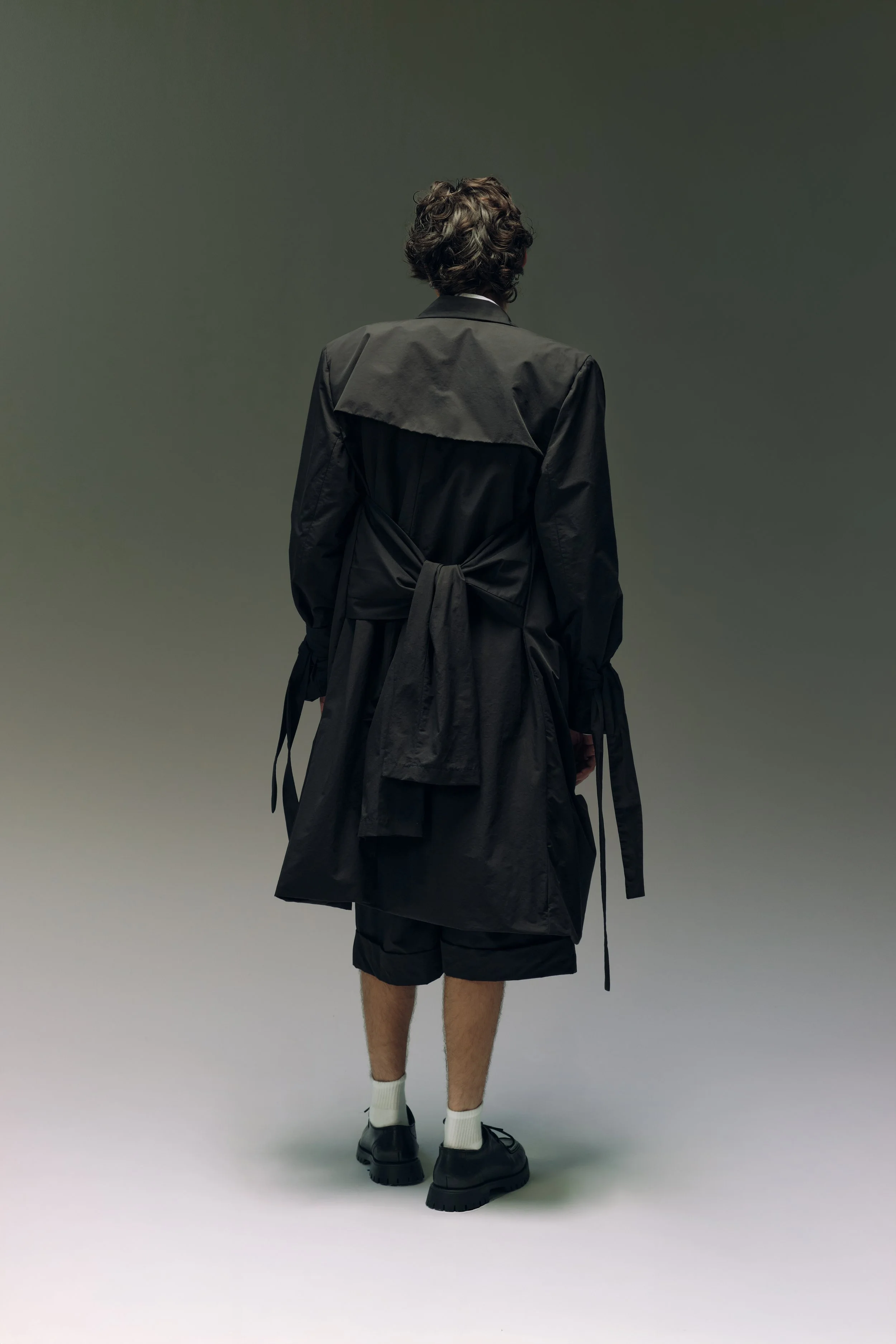 Persona vista de espaldas, usando abrigo negro, pantalones cortos y zapatos negros, en fondo gris