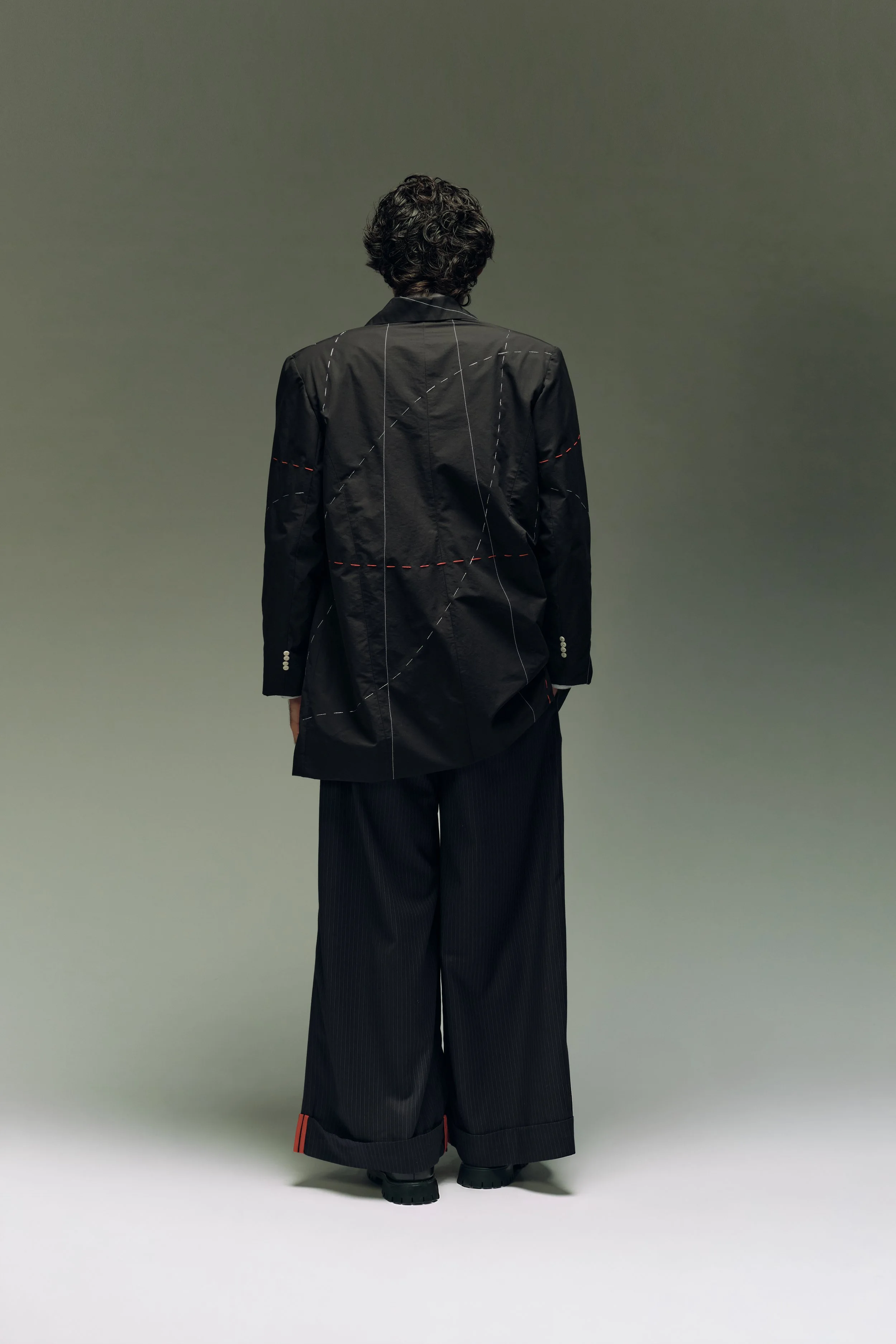 Persona de espaldas con ropa negra, chaqueta con costuras y pantalones anchos, fondo gris liso.