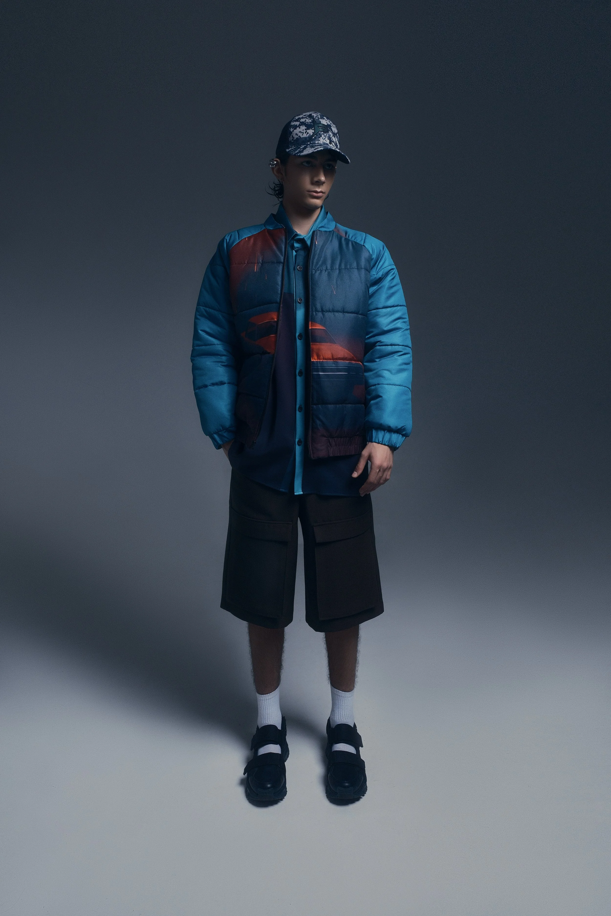 Joven con gorra, chaqueta azul con diseño, pantalones cortos negros, calcetas blancas y sandalias negras, en fondo gris oscuro.