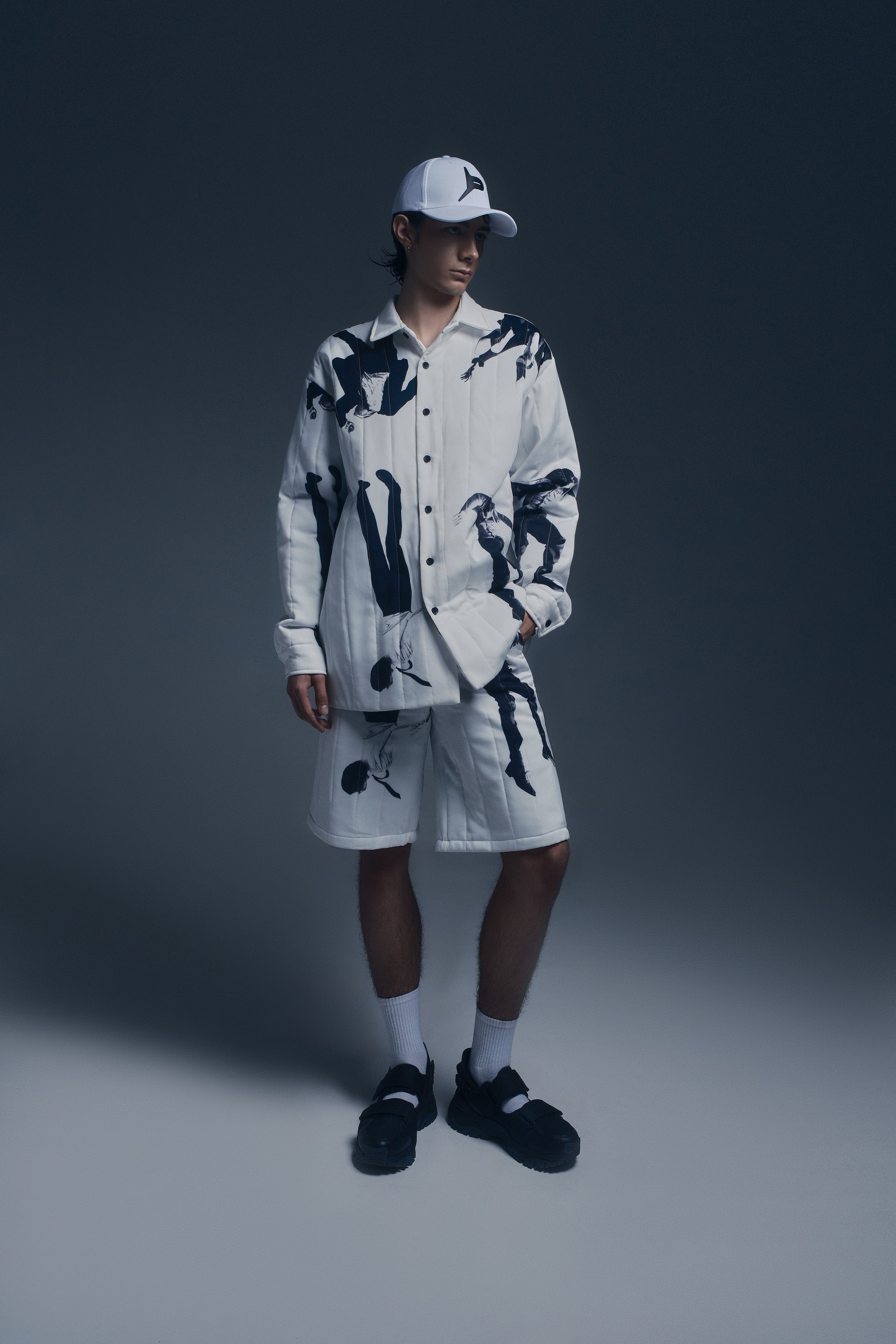 Joven vestido con ropa deportiva blanca con estampado oscuro, gorra blanca, calcetas blancas y sandalias negras, en un fondo gris oscuro.