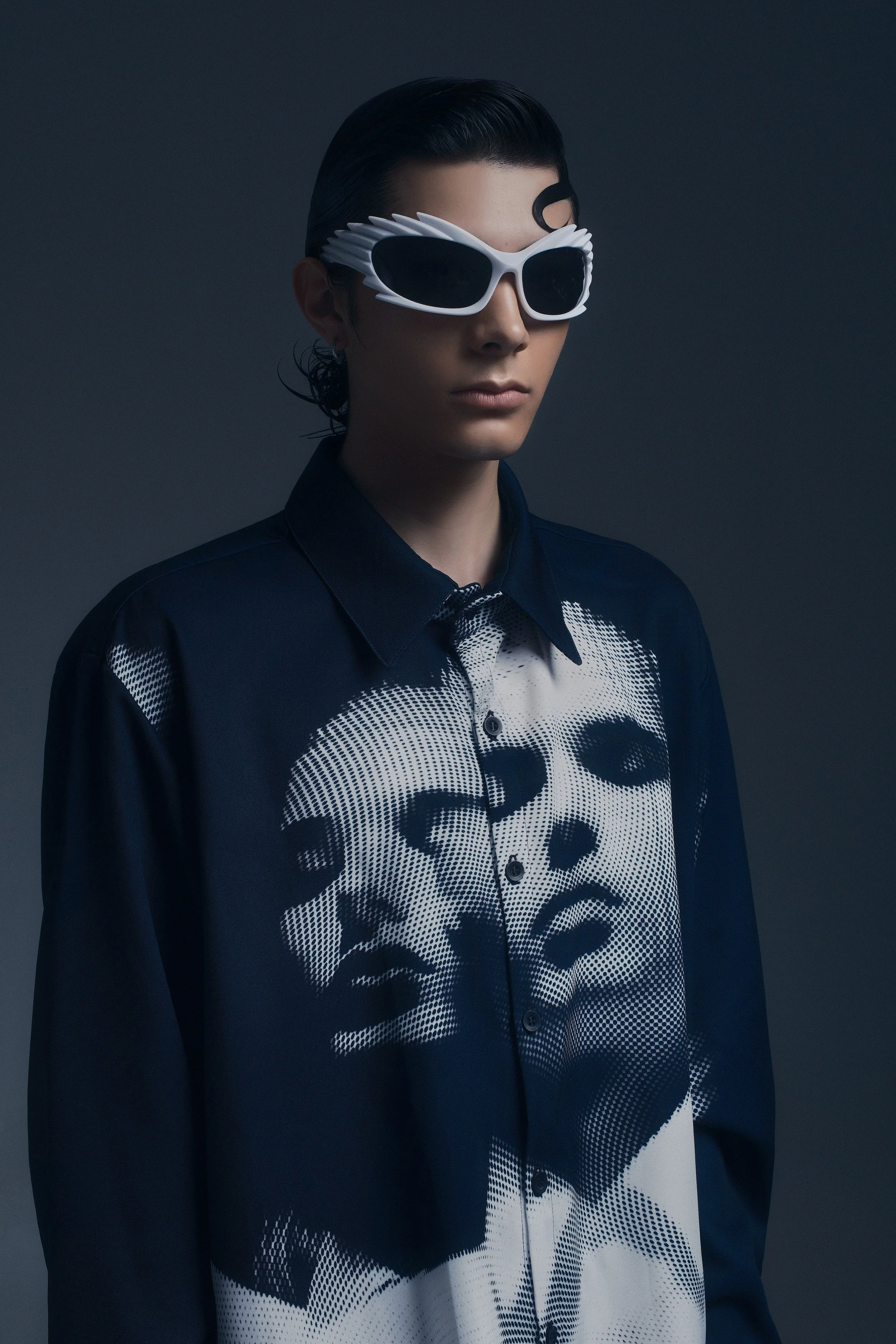 Mujer con gafas blancas con diseño futurista y camisa con gráficos de retratos de rostros humanos en tonos oscuros y claros en un fondo gris oscuro.