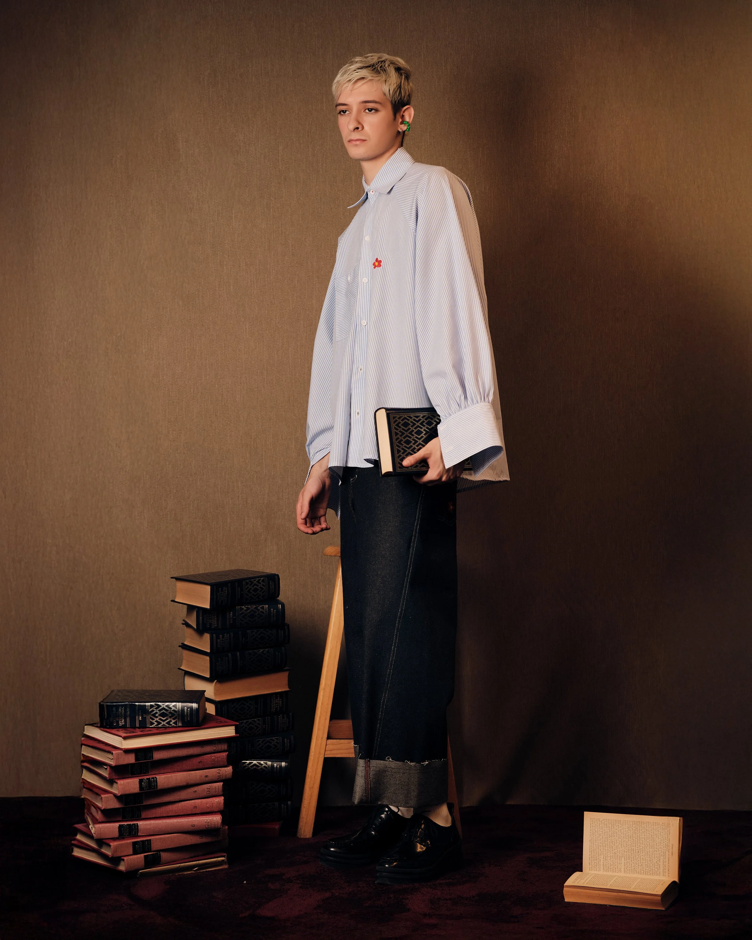 Persona joven con cabello rubio cortado a la moda, sosteniendo un libro, rodeado de pilas de libros en un fondo marrón, vestido con una camisa de rayas y pantalones anchos de mezclilla, en un entorno de estudio o biblioteca.