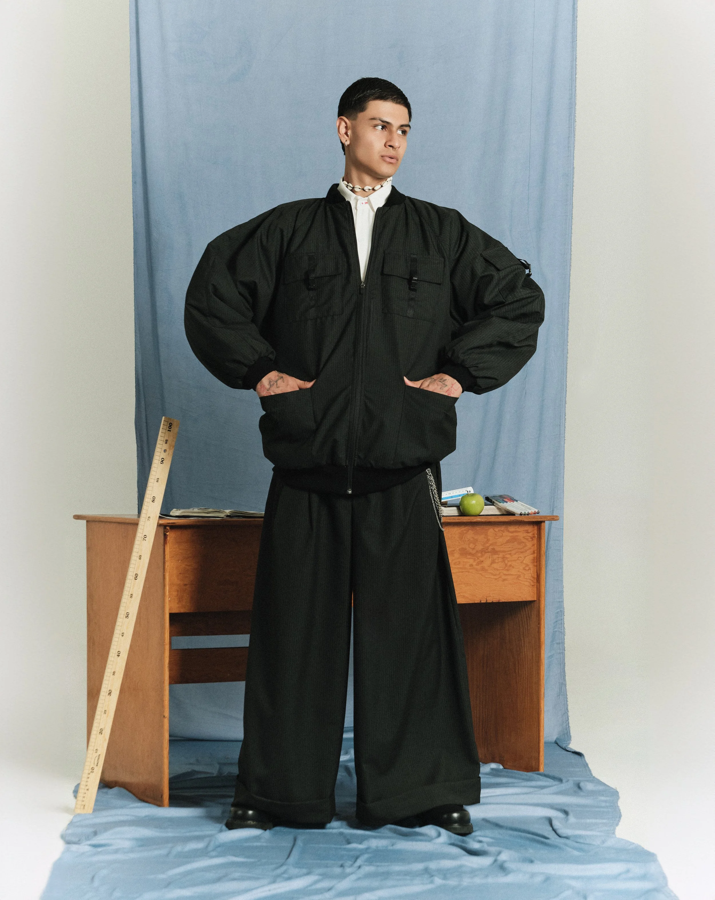 Persona joven con cabello corto, camiseta blanca y chaqueta negra grande, de pie en un fondo azul con muebles y objetos como una regla, una regla de cartulina, una manzana, algunos libros y unos útiles escolares sobre una mesa.