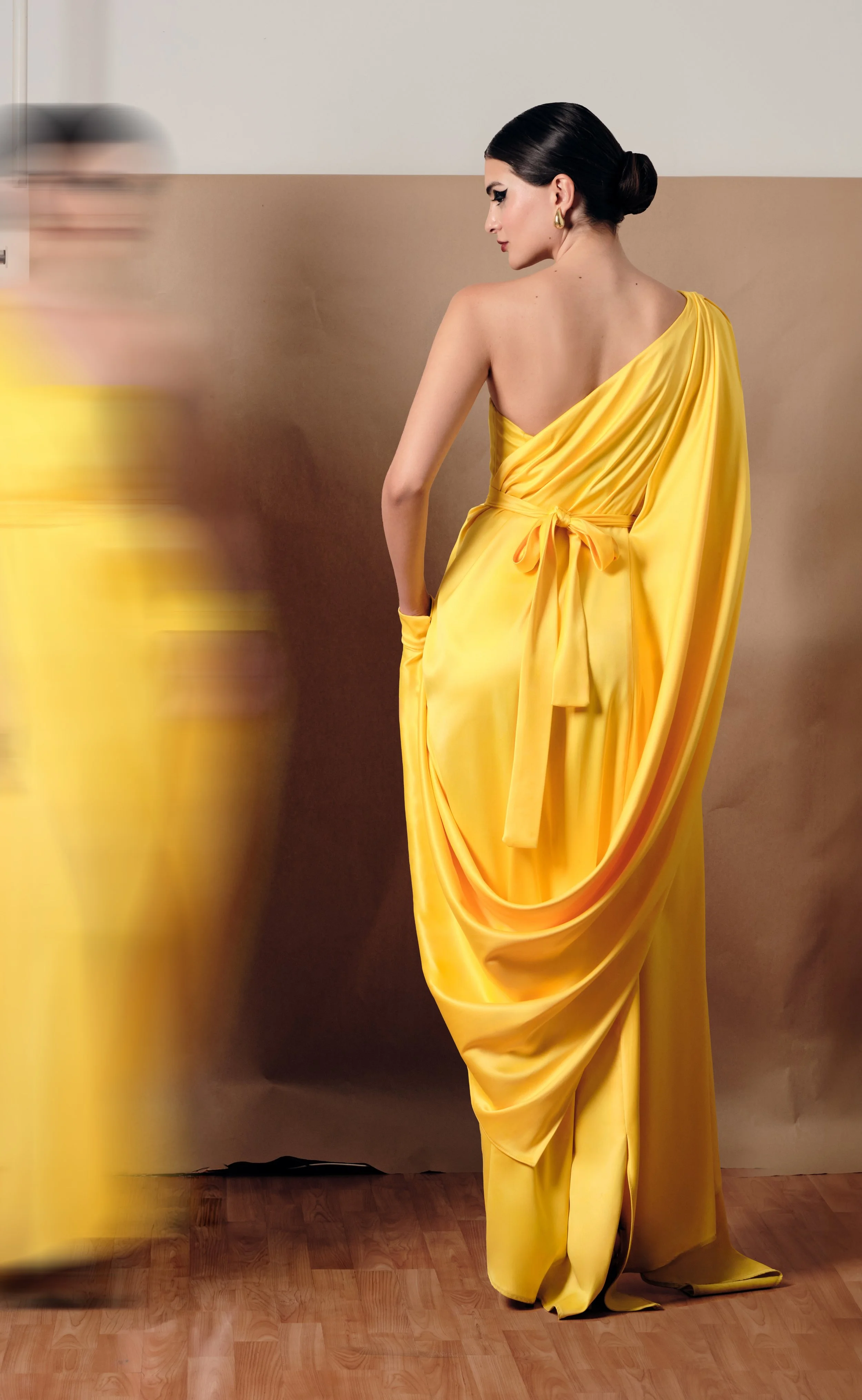 Mujer con vestido amarillo brillante, peinado elegante en cofia, usando pendientes dorados, en un fondo neutro, con movimiento borroso en la izquierda.