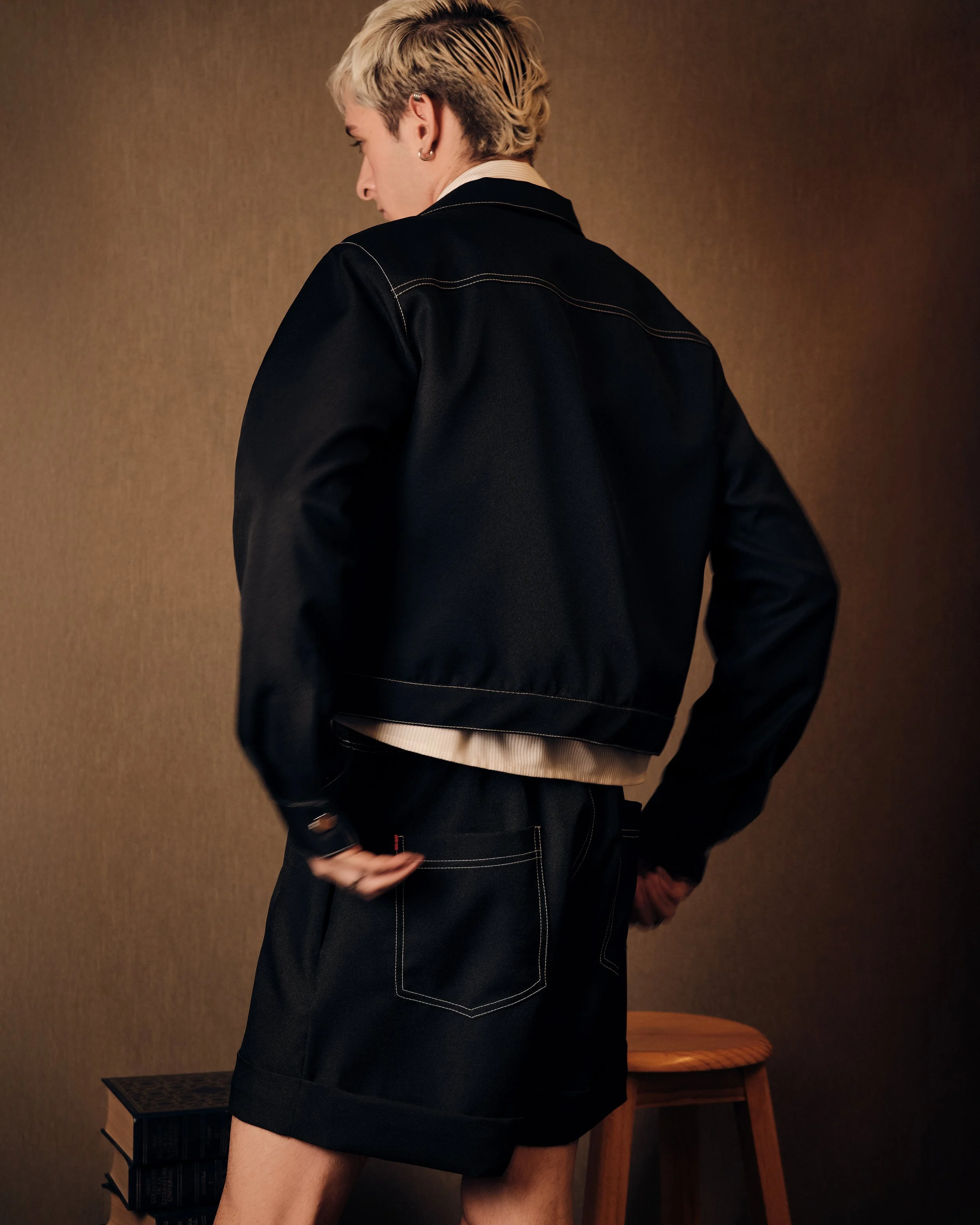 Persona joven con cabello rubio platinado, vista de espaldas, usando chaqueta negra y pantalones cortos negros con costuras blancas, en un fondo marrón.