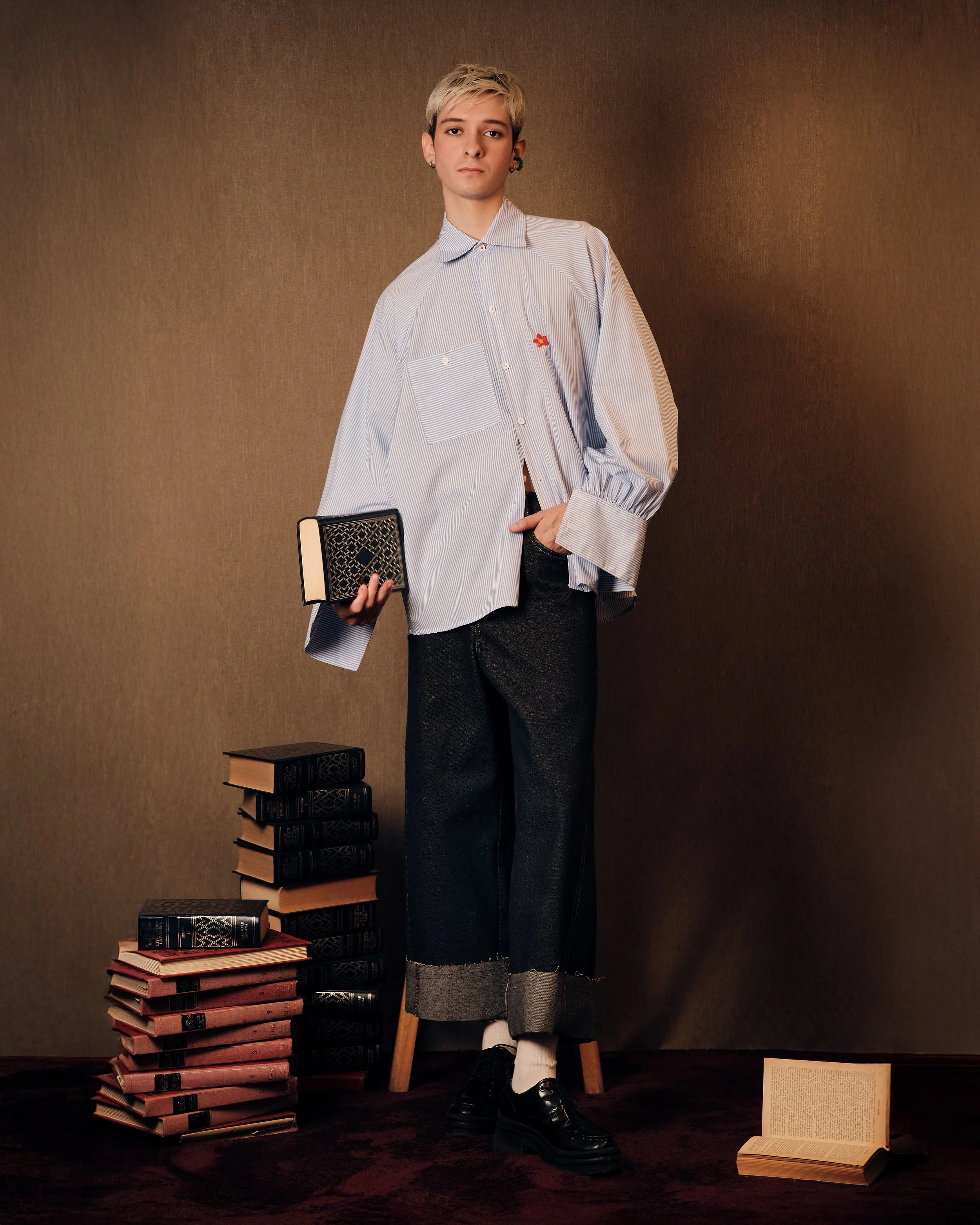Joven con cabello rubio corto, ropa casual con camisa de rayas, sosteniendo libro en la mano, rodeado de pilas de libros en el suelo y fondo de tela marrón.
