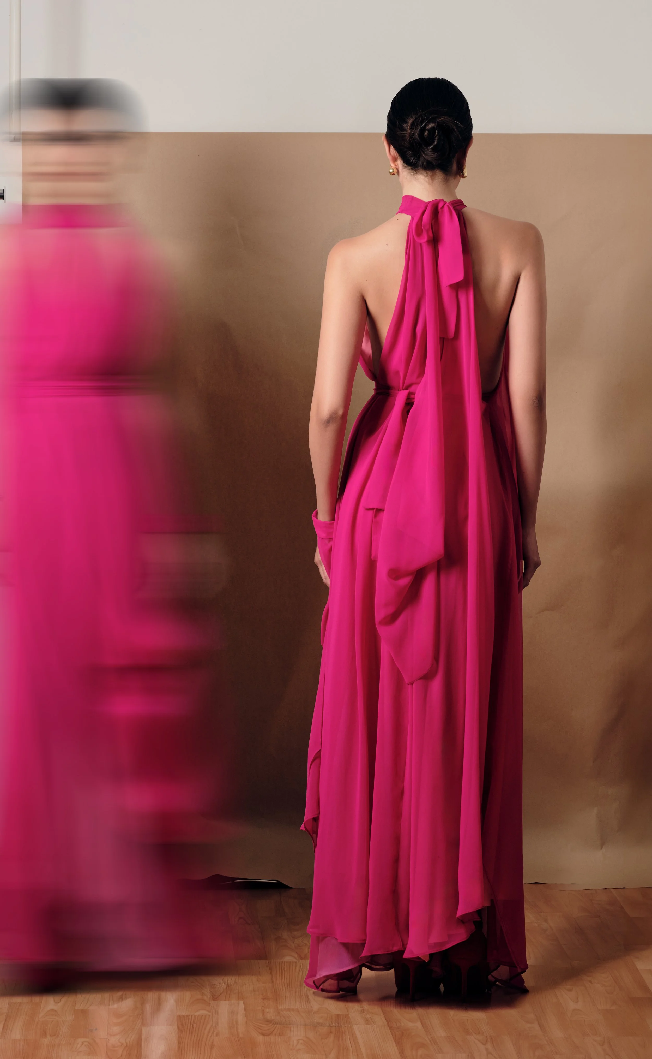 Mujer vista desde atrás con vestido largo rosa, cabello recogido y pendientes dorados, en un fondo de pared beige y blanca, con piso de madera.