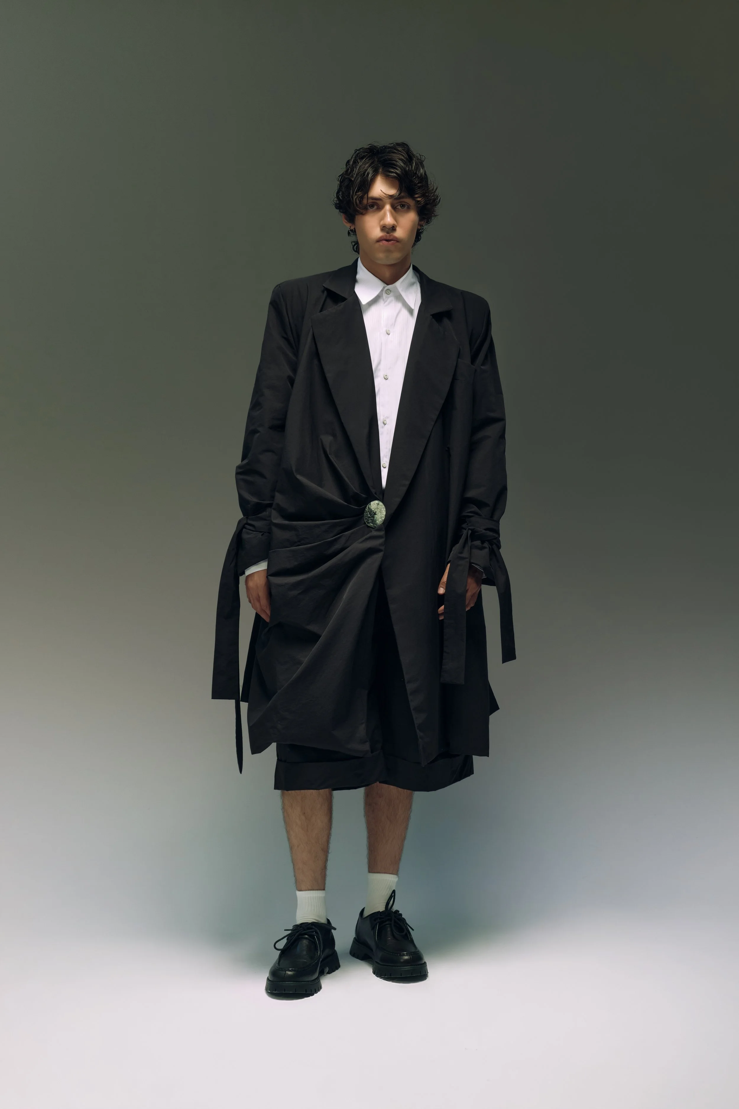 Joven con cabello rizado, usando un conjunto de ropa negra, con una camisa blanca y zapatos negros, posando en un fondo gris.