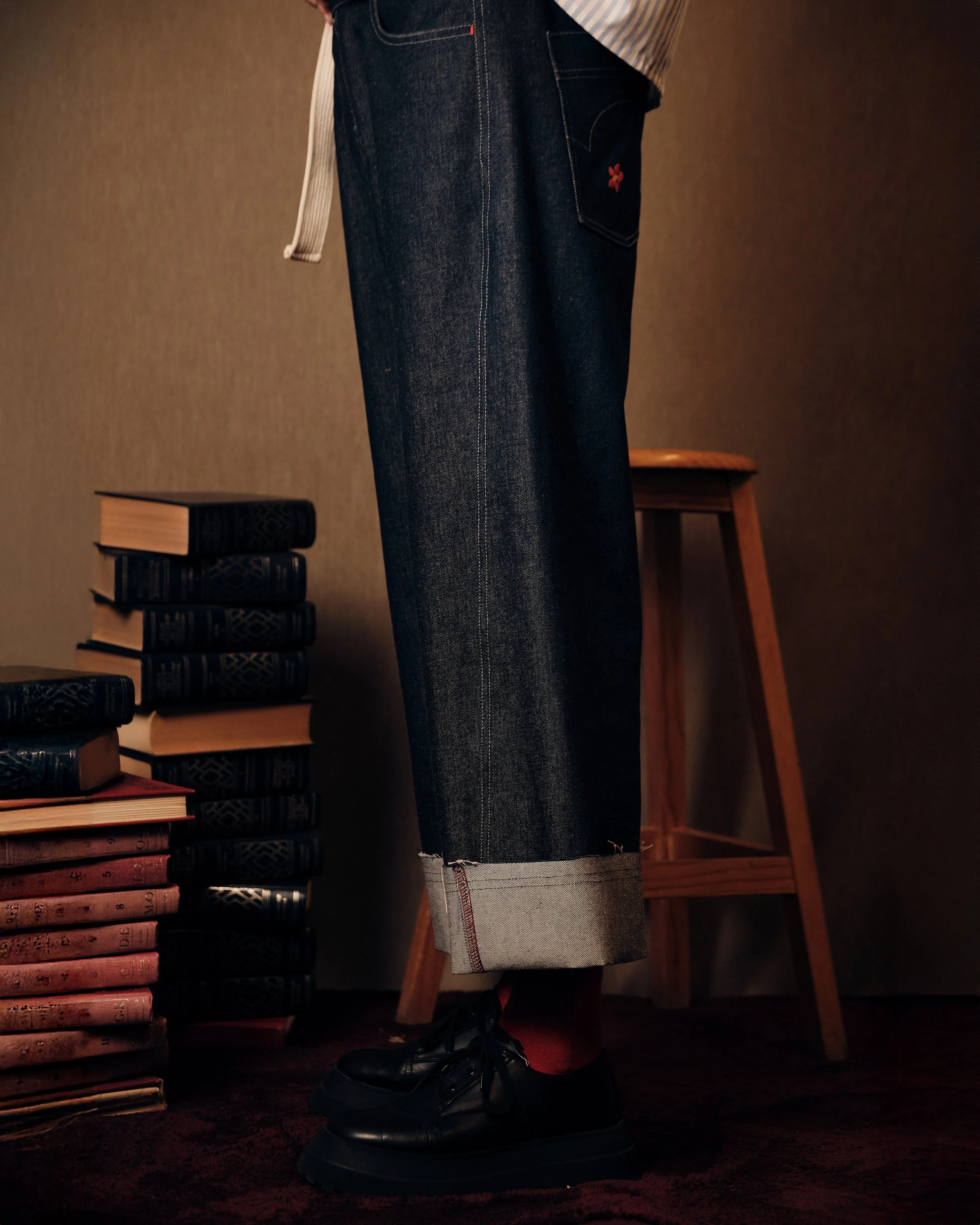 Persona usando pantalones de mezclilla enrollados, zapatos negros y calcetas rojas, rodeada de libros en una habitación